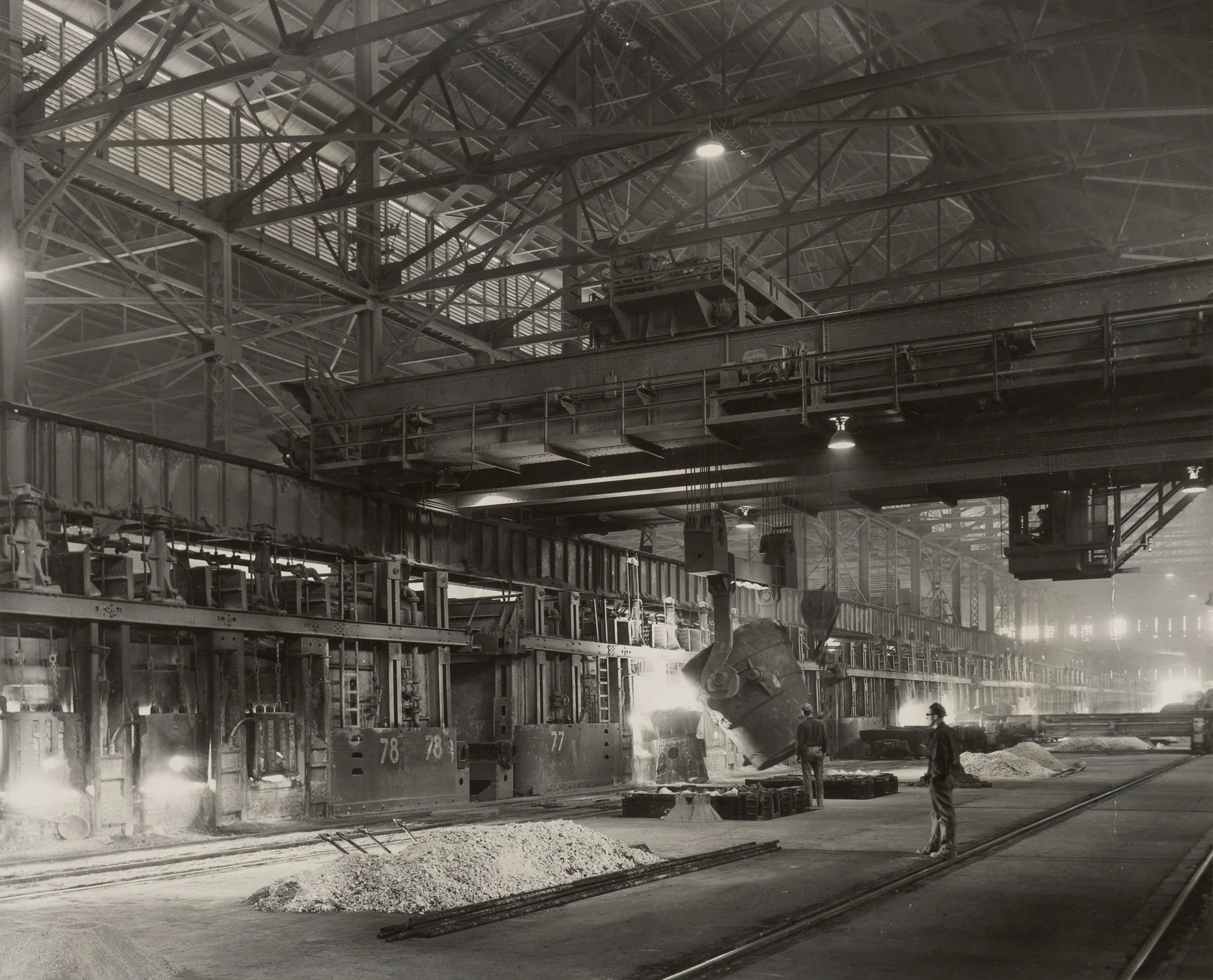 Large open hearth steel mill with overhead crane pouring molten iron into a furnace; workers stand nearby.