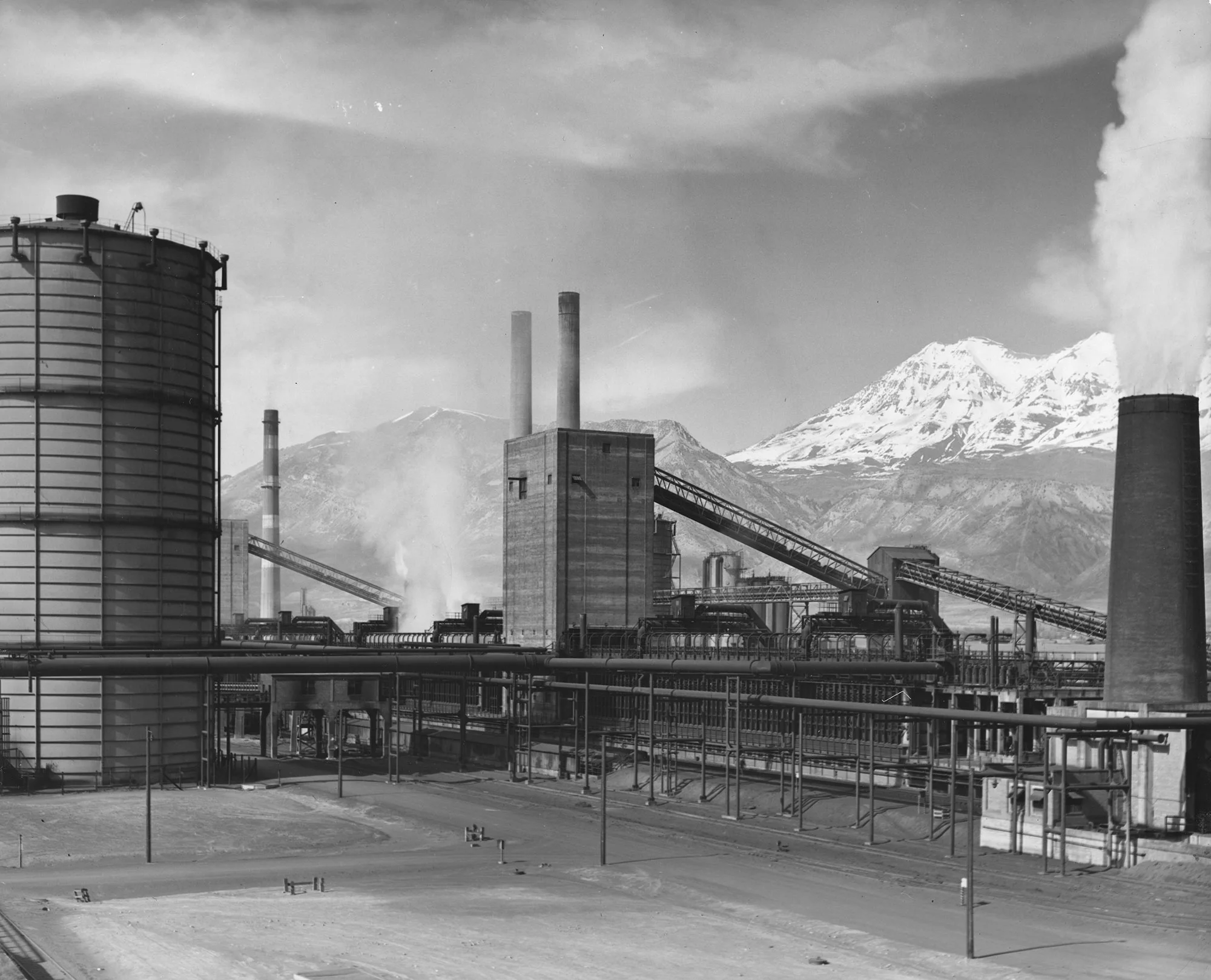Coke ovens and smokestacks at Geneva Steel Plant in Utah with snowy mountains in the background and steam rising from stacks.
