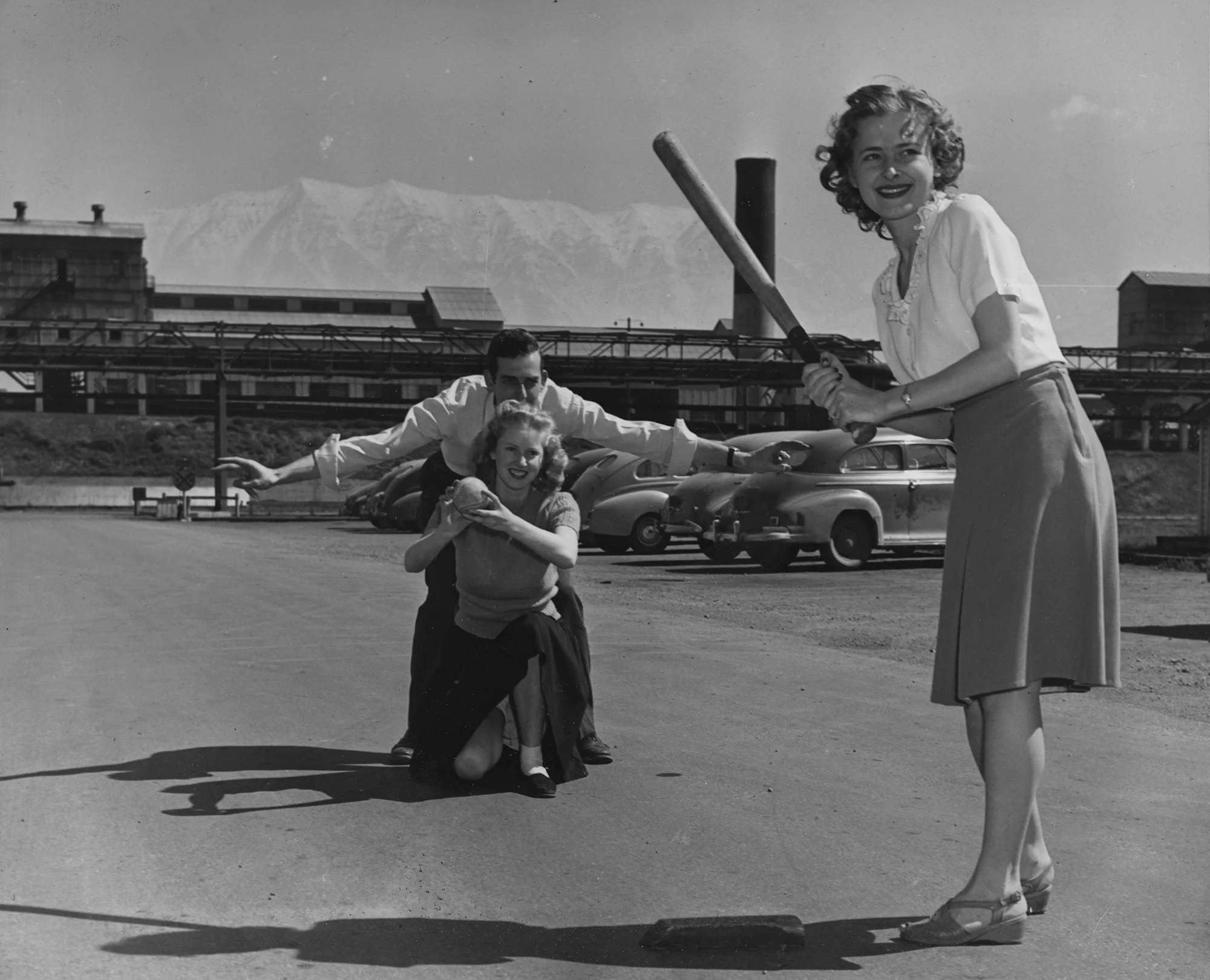 Three adults play baseball in a steel mill parking lot with vintage cars and snow-capped mountains in the background.