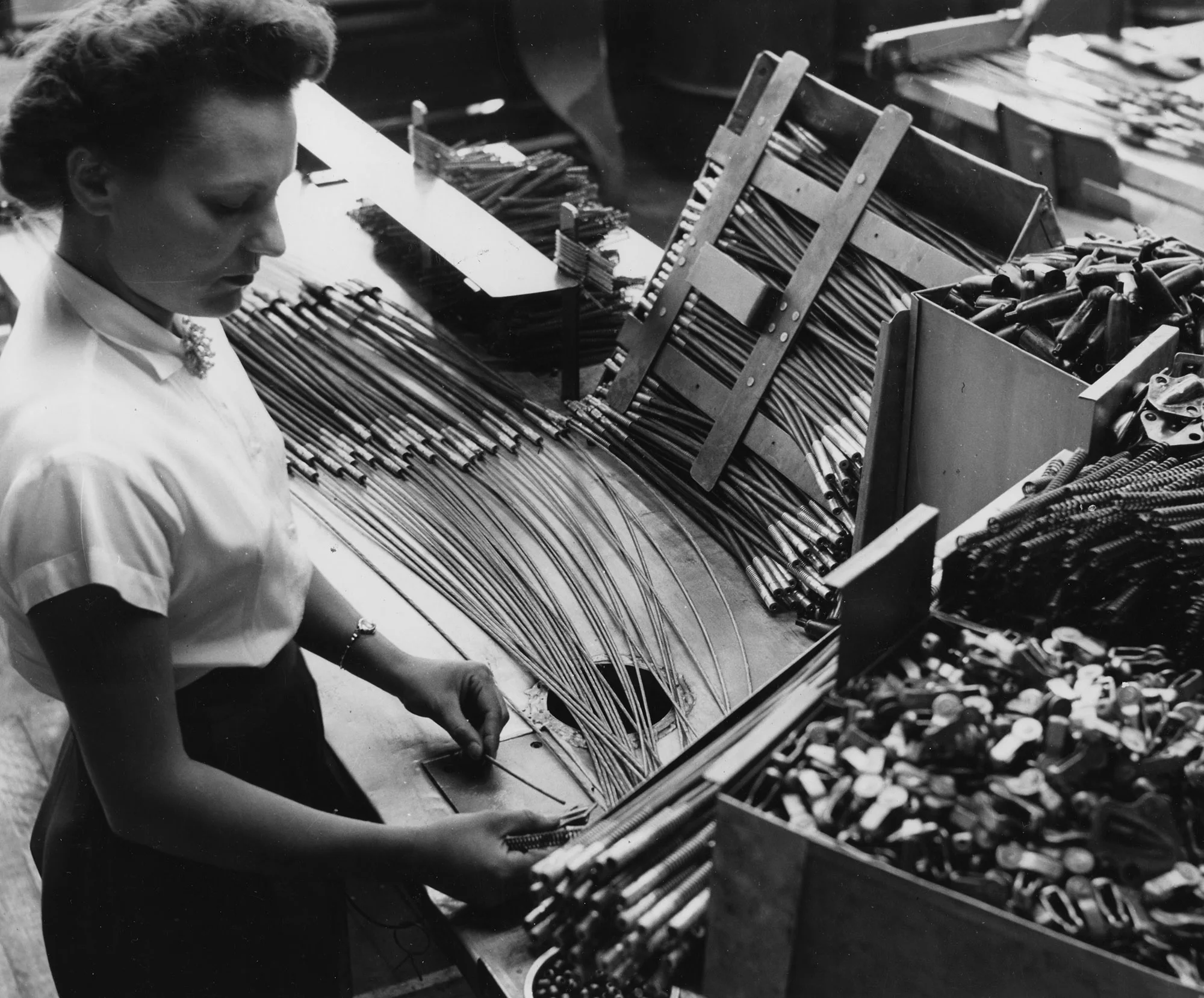 Factory worker organizes metal rods and components on an assembly table surrounded by bins.