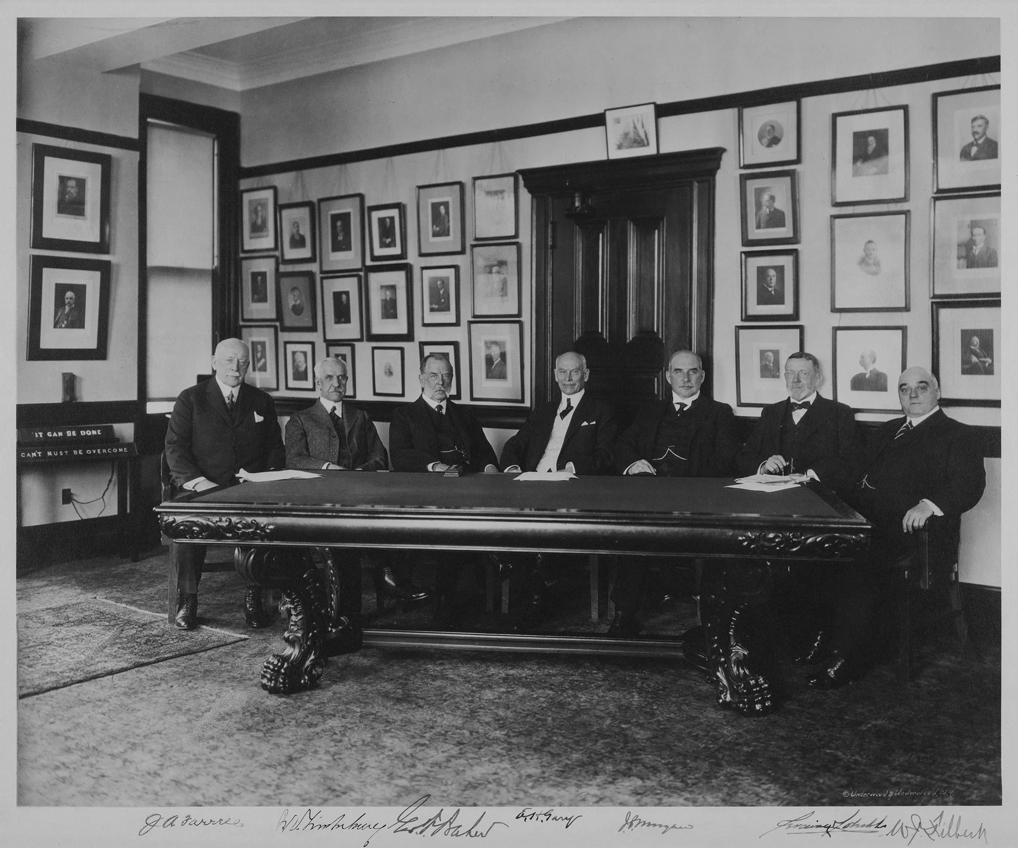 Seven executives sit at a long ornate table in a portrait-lined boardroom, posing for a formal group photo.