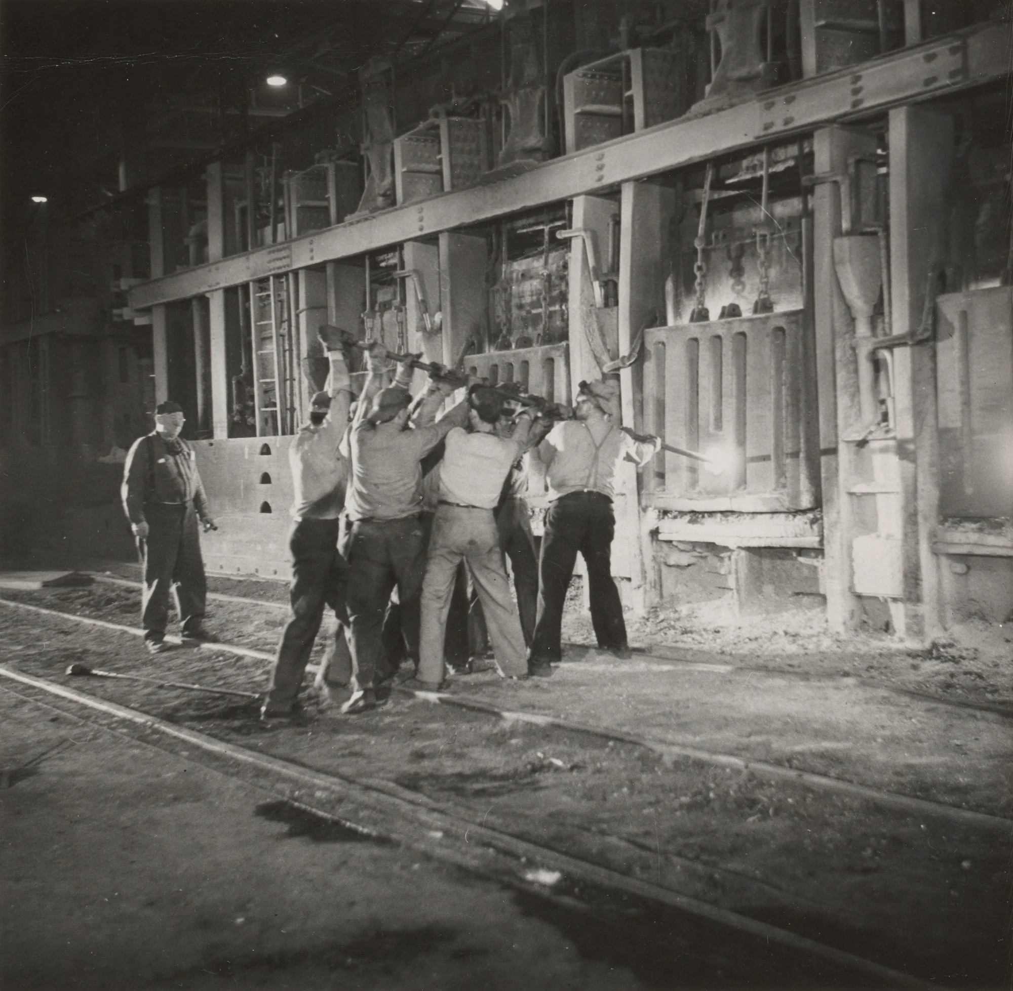 Team of workers uses long rod to tap open hearth furnace at Gary Steel Works during steelmaking process.