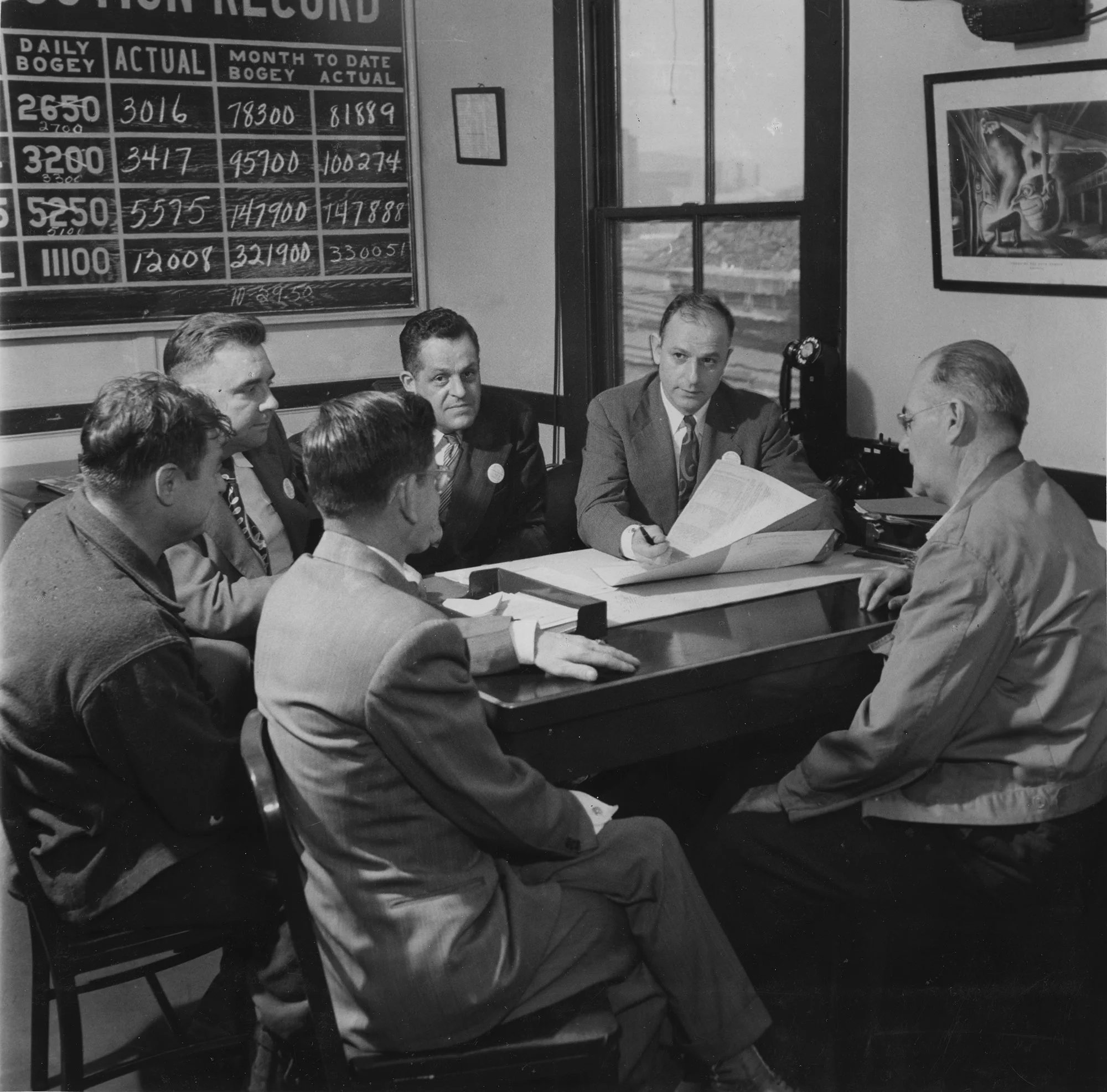 Six men in suits and workwear sit around a table reviewing production data in an office with a steel output board.