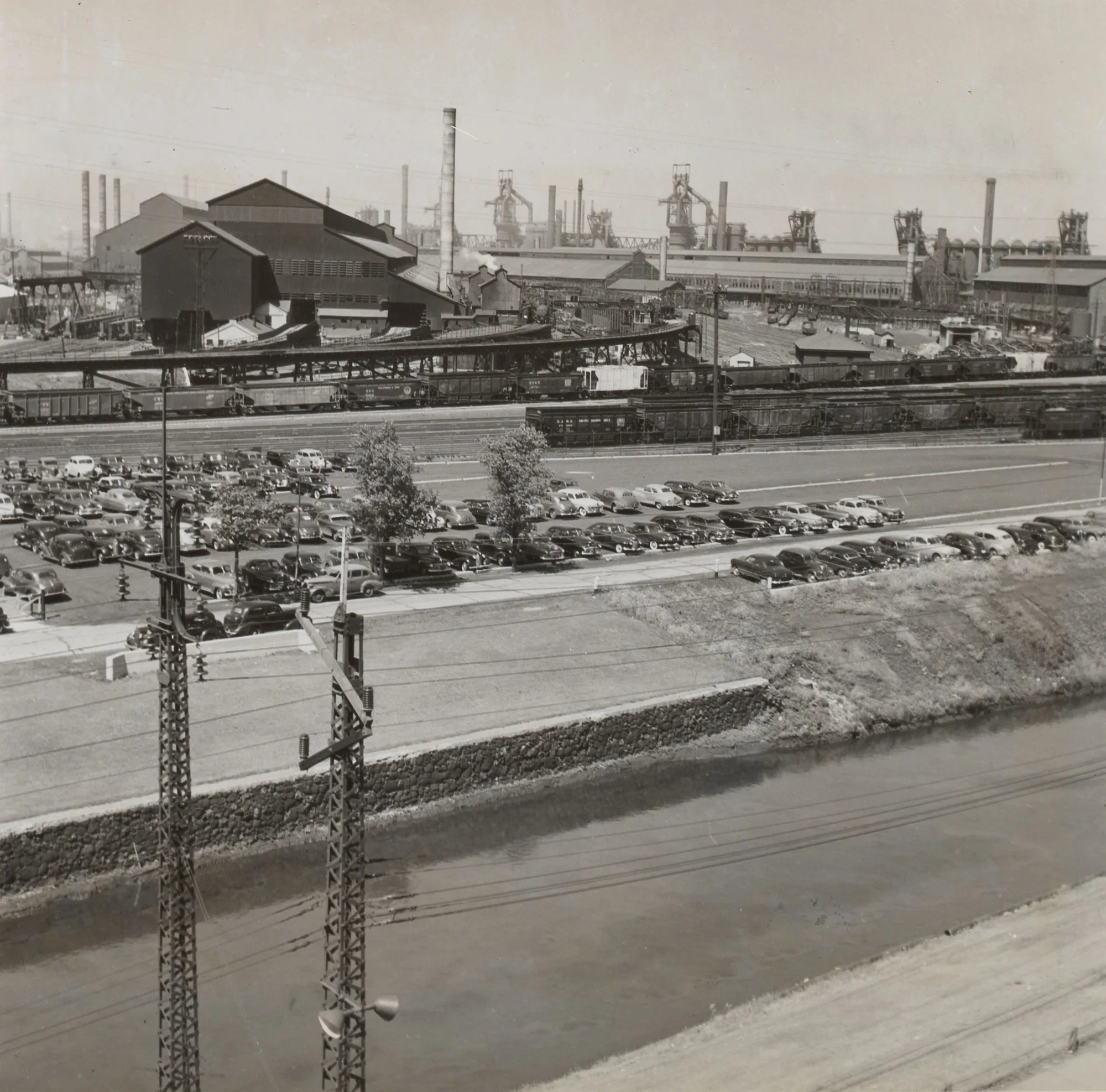 Industrial steel plant complex with blast furnaces, railcars, and a full parking lot across from a canal or drainage ditch.