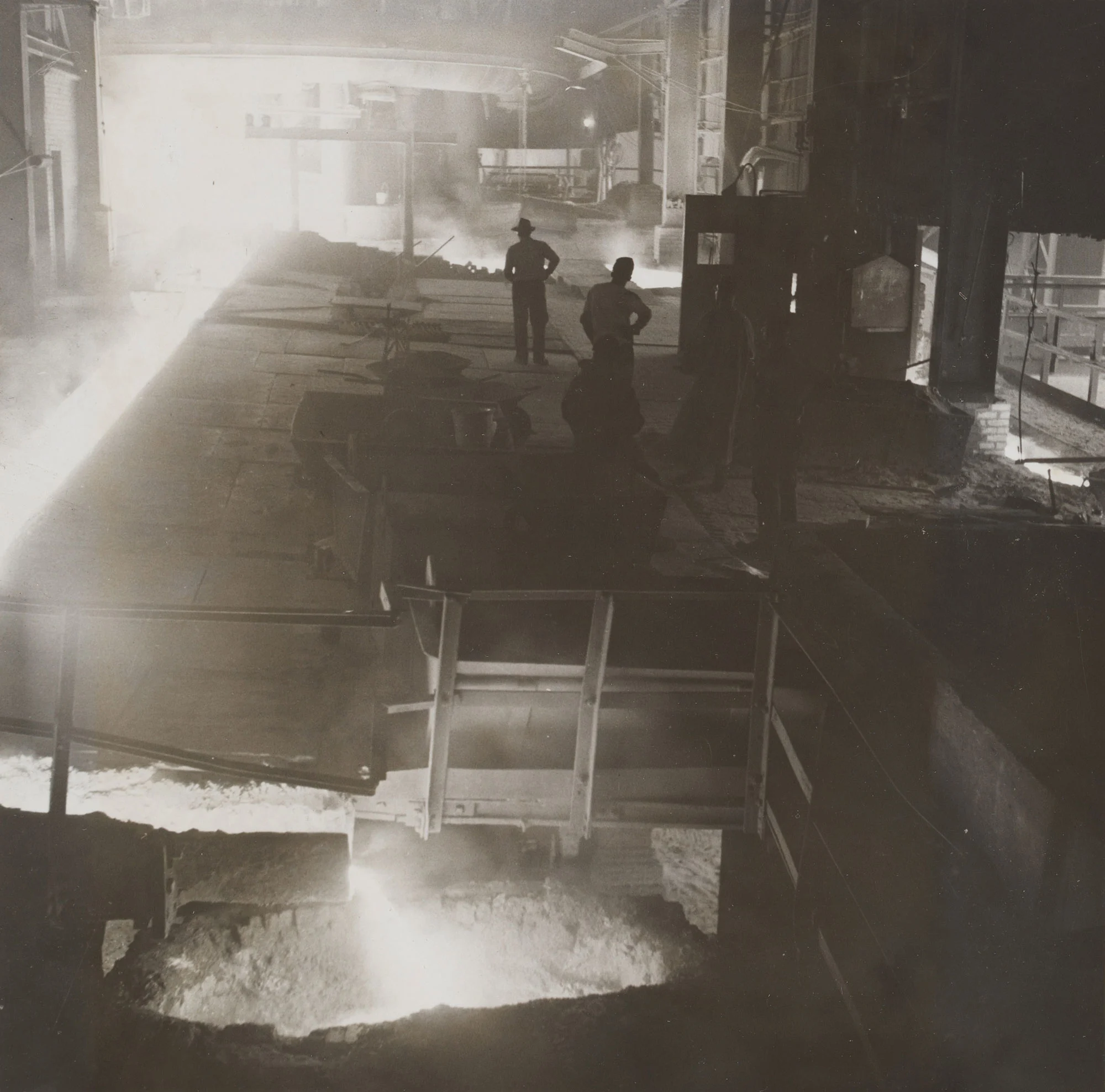 Workers observe molten metal pouring into a pit inside a dimly lit steel mill filled with steam, smoke, and intense light.