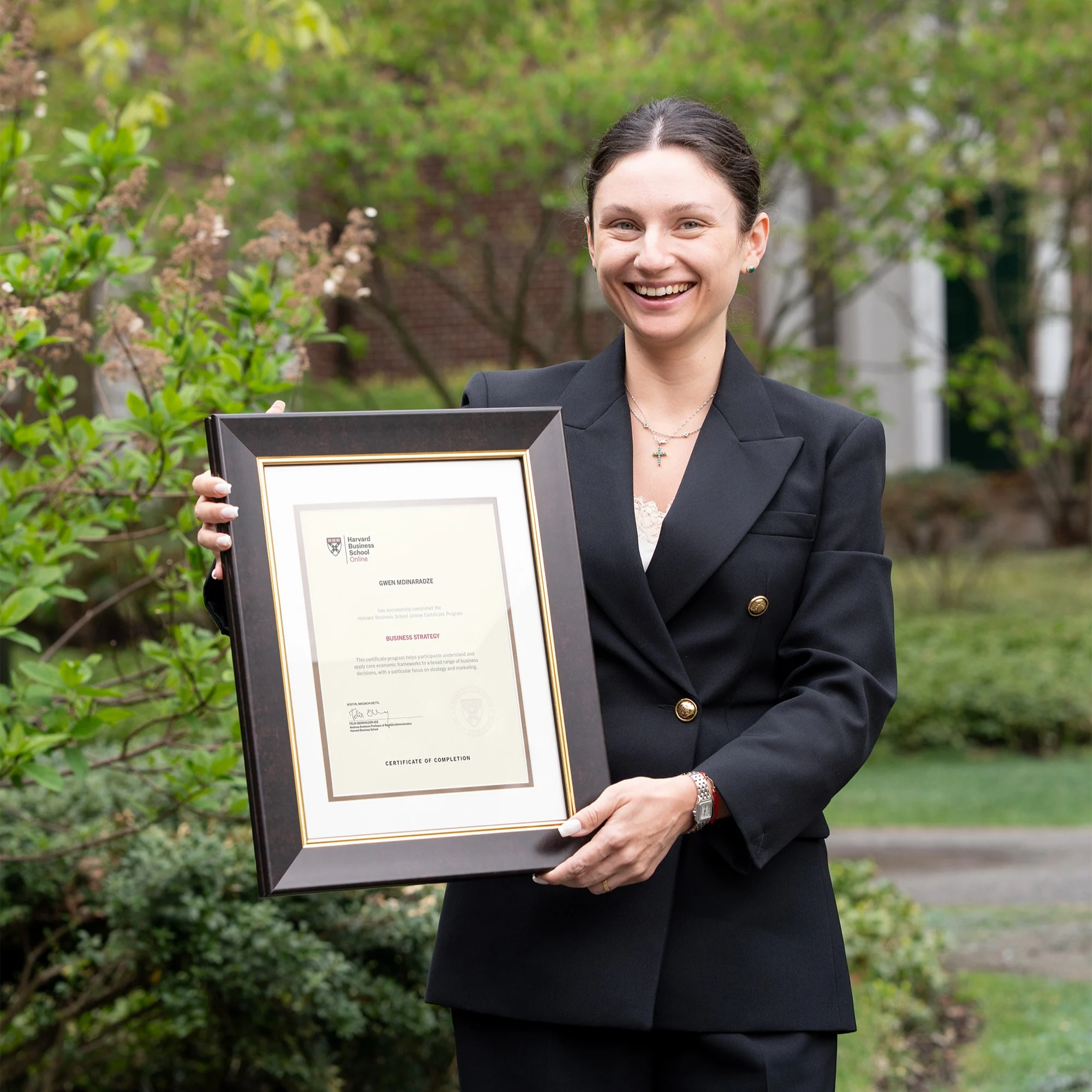 HBS Online community organizer in black suit smiling and holding a framed Harvard certificate outdoors.
