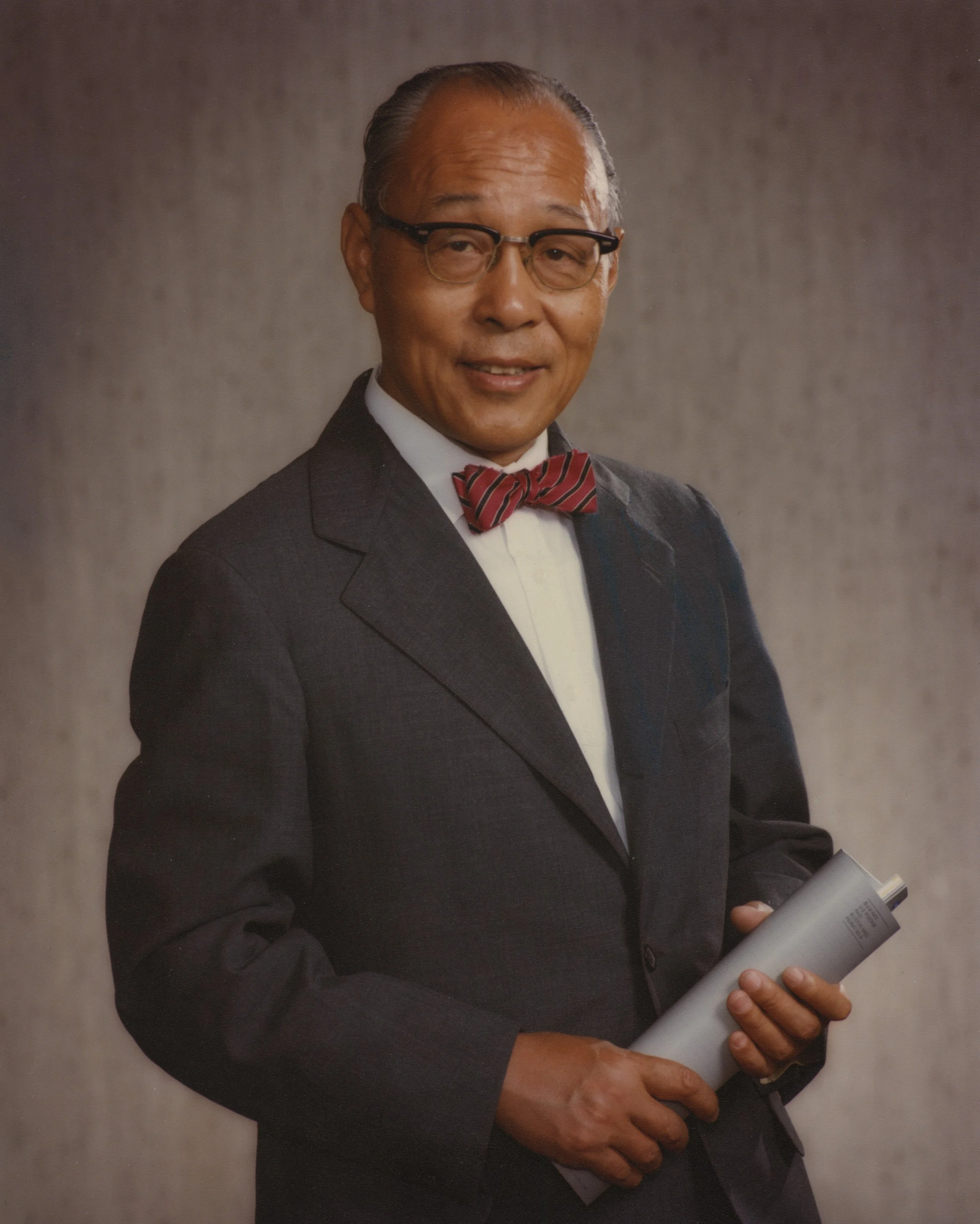 A man with glasses in a suit with a bow tie holding a cylindrically folded document