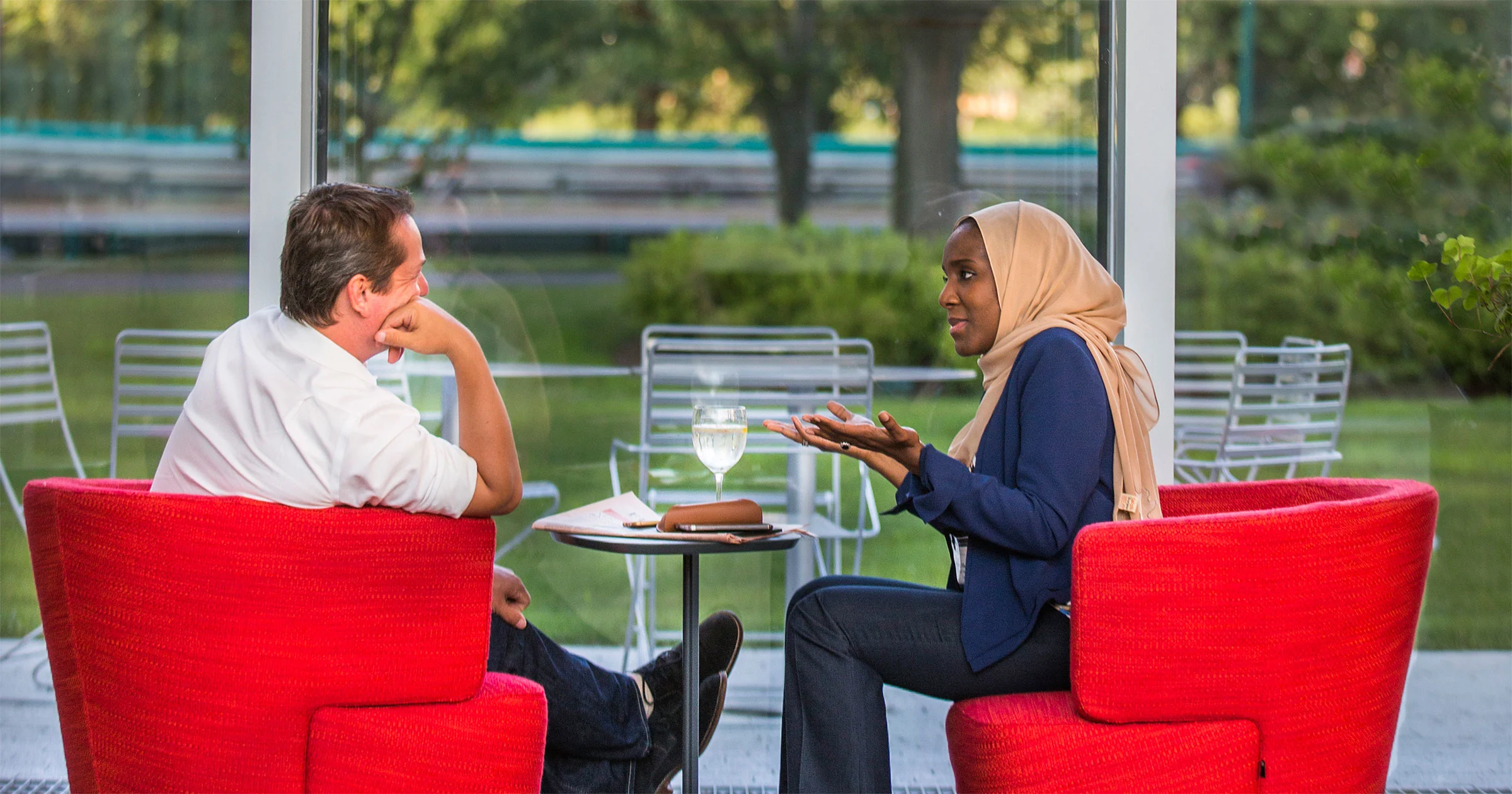 Male and female executive education participants sit in red chairs by a window, engaged in conversation. The woman, wearing a hijab, gestures while speaking