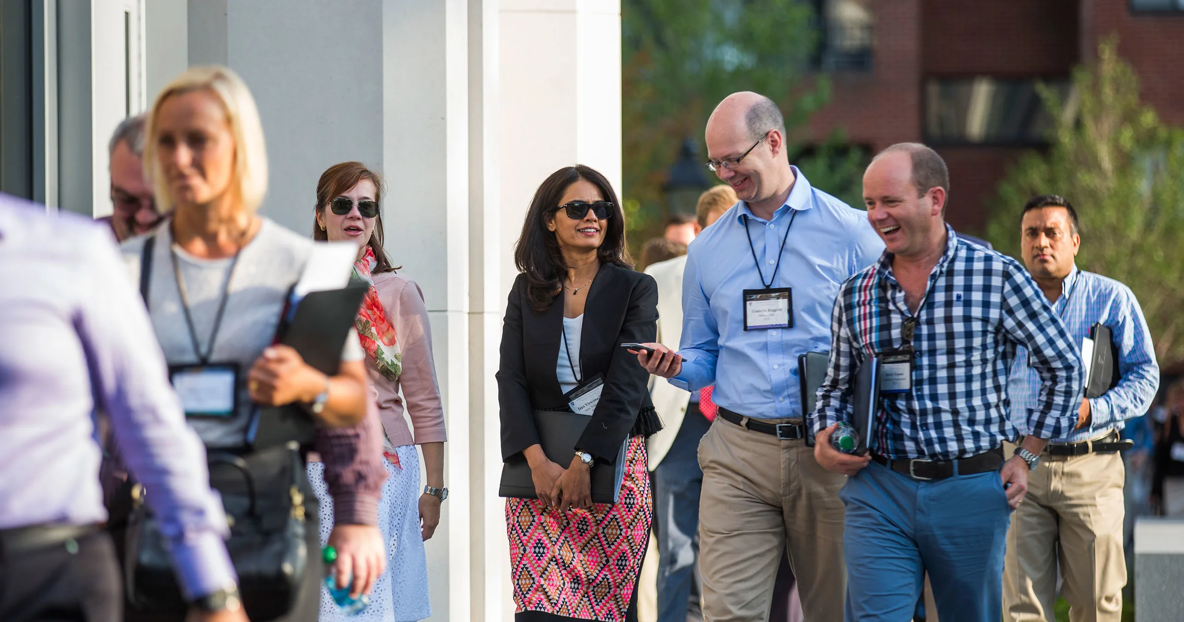 A group of executives, wearing business casual attire and name badges, walk together on HBS campus, engaged in conversation and laughter