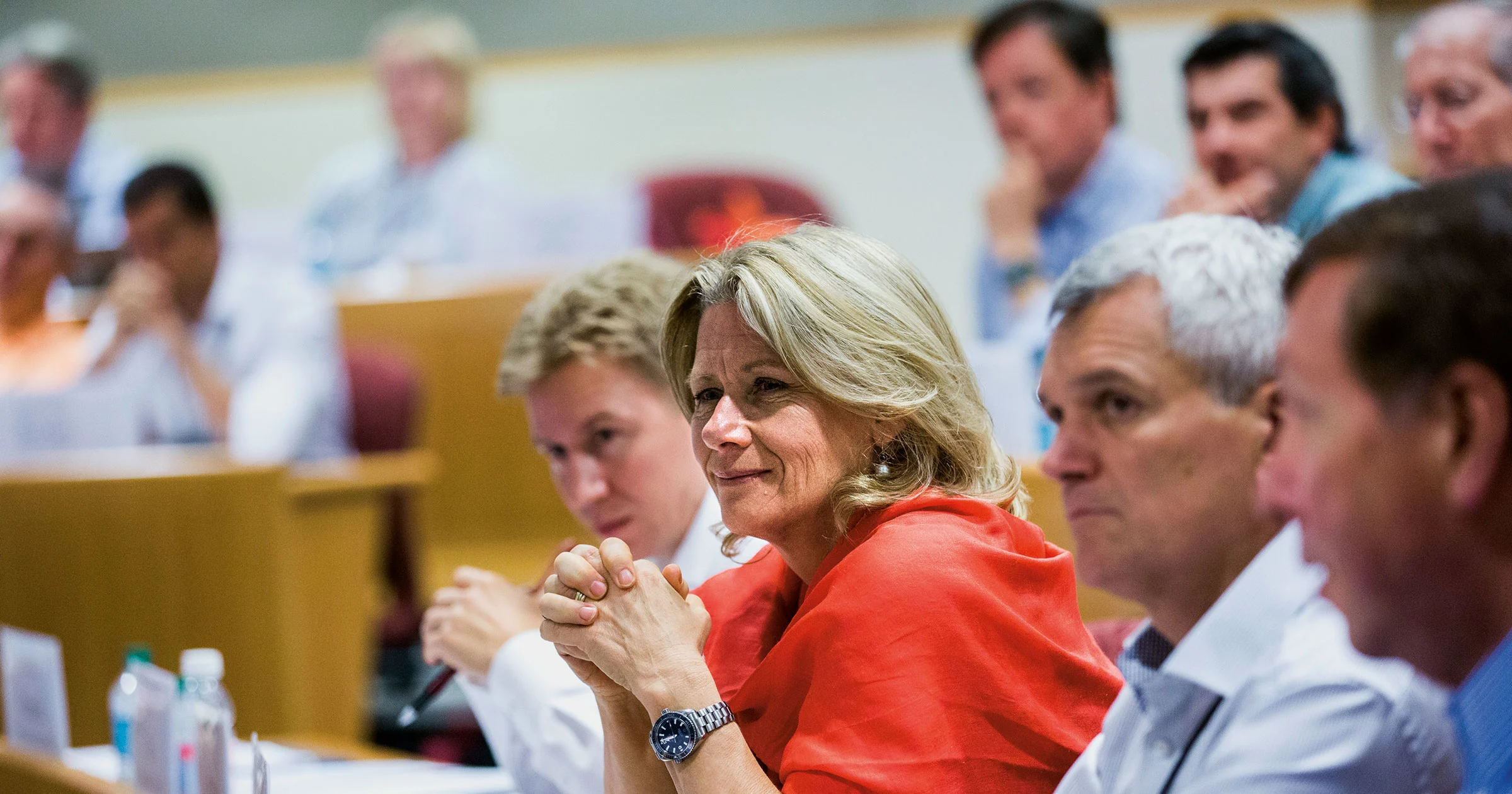 A group of executives attentively listening in a lecture hall, with a woman in a red shawl smiling