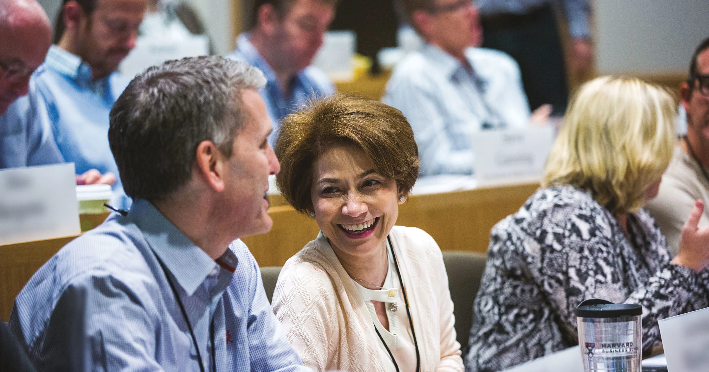 Executives team up in classroom discussion, with a smiling woman in focus. Harvard Business School logo visible