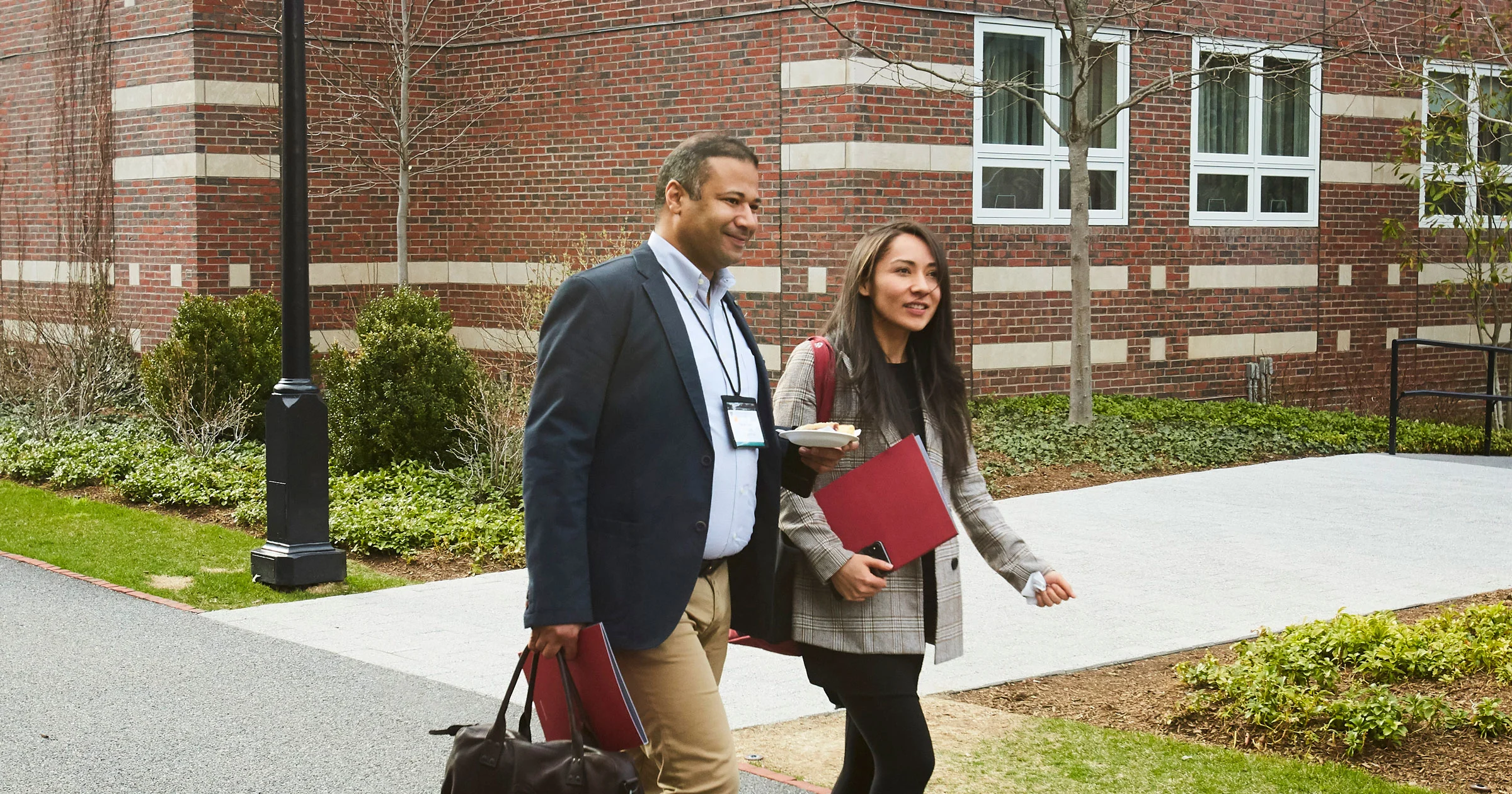 Two executives walk and chat on the HBS campus, carrying folders and bags, dressed in business casual attire