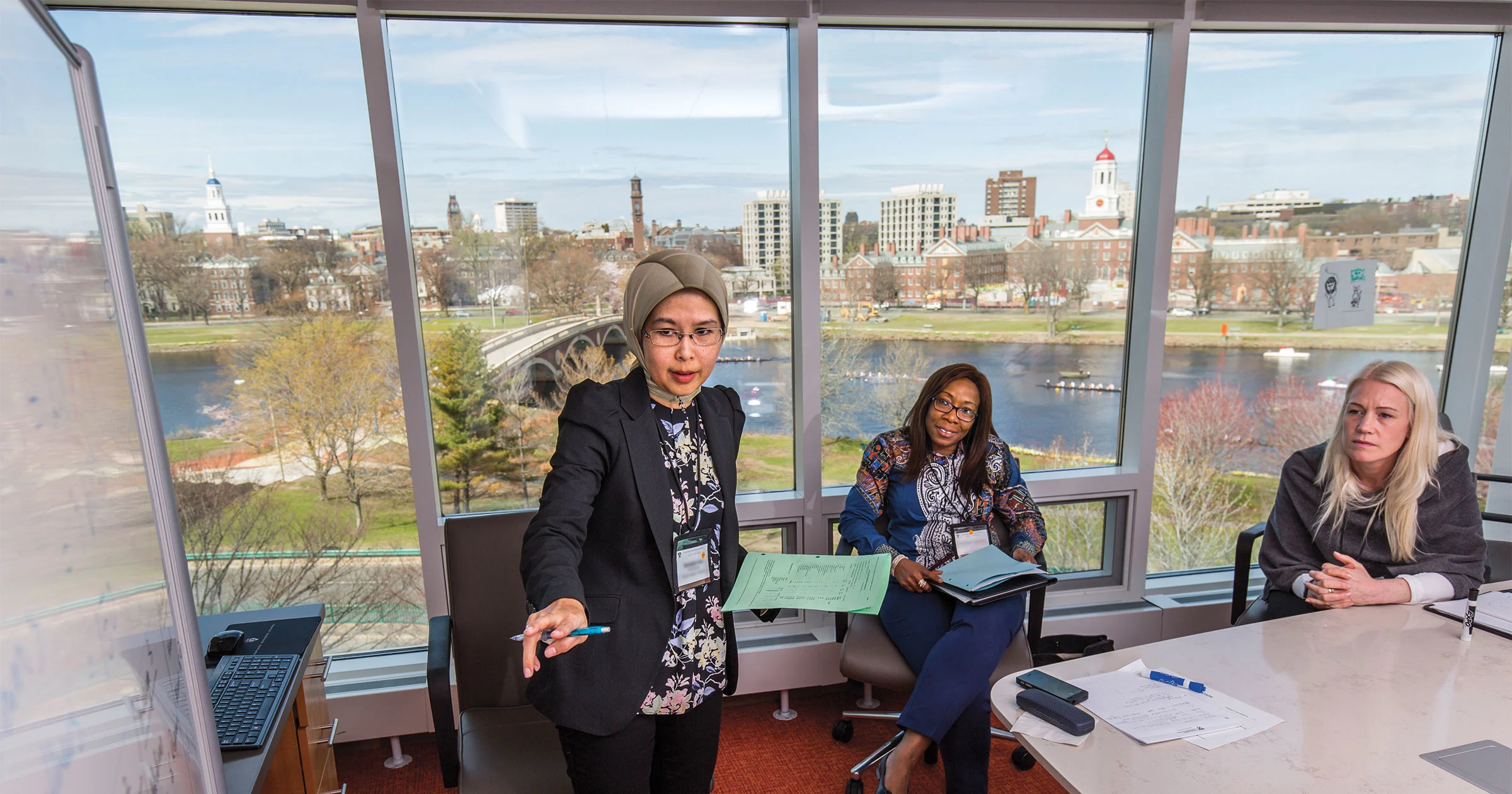 A female executive education participant leads a discussion in a conference room with Charles river view behind her