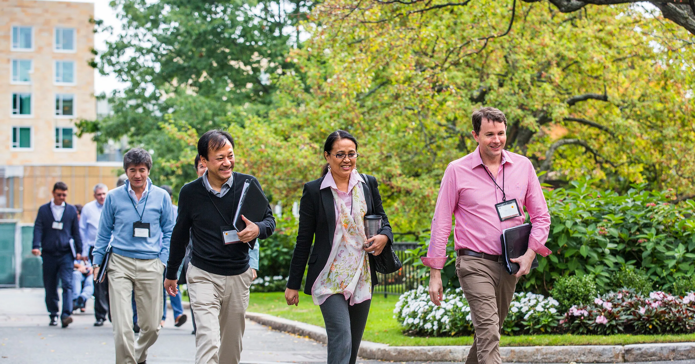 Group of executives walking outdoors on a green HBS campus, engaged in conversation and carrying notebooks