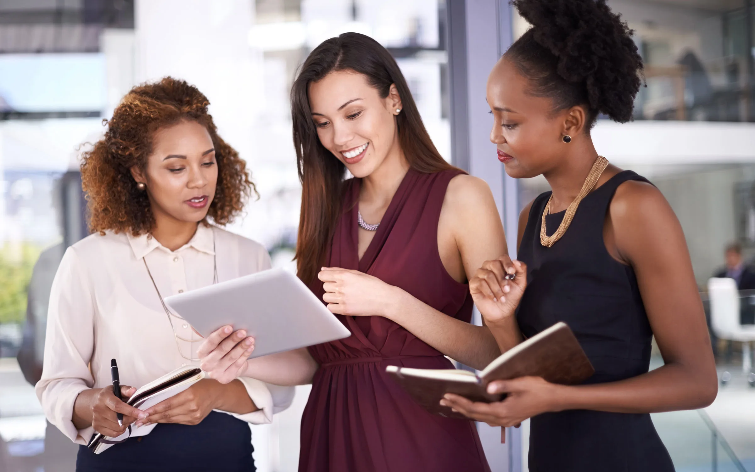 Three women of color executives standing discussion, reviewing a tablet and taking notes with notebooks
