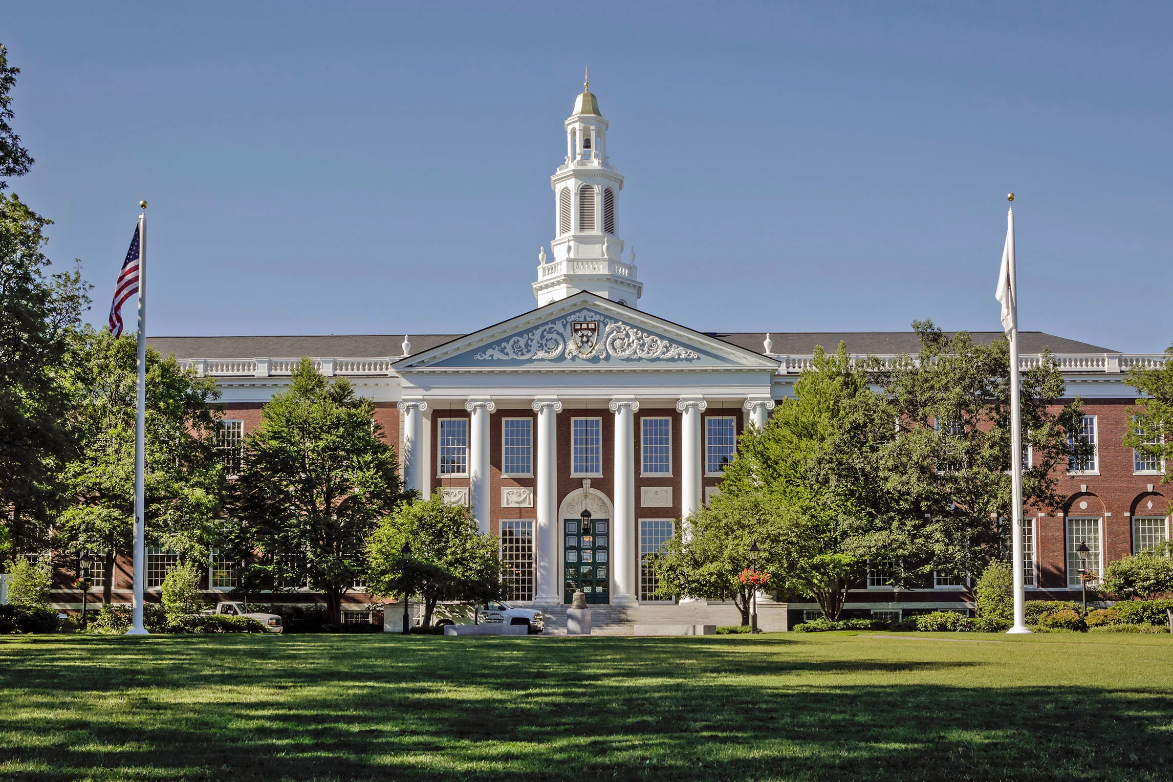 Front view of Baker Library with white columns, a central cupola, flagpoles, trees, and a green lawn under a clear blue sky.