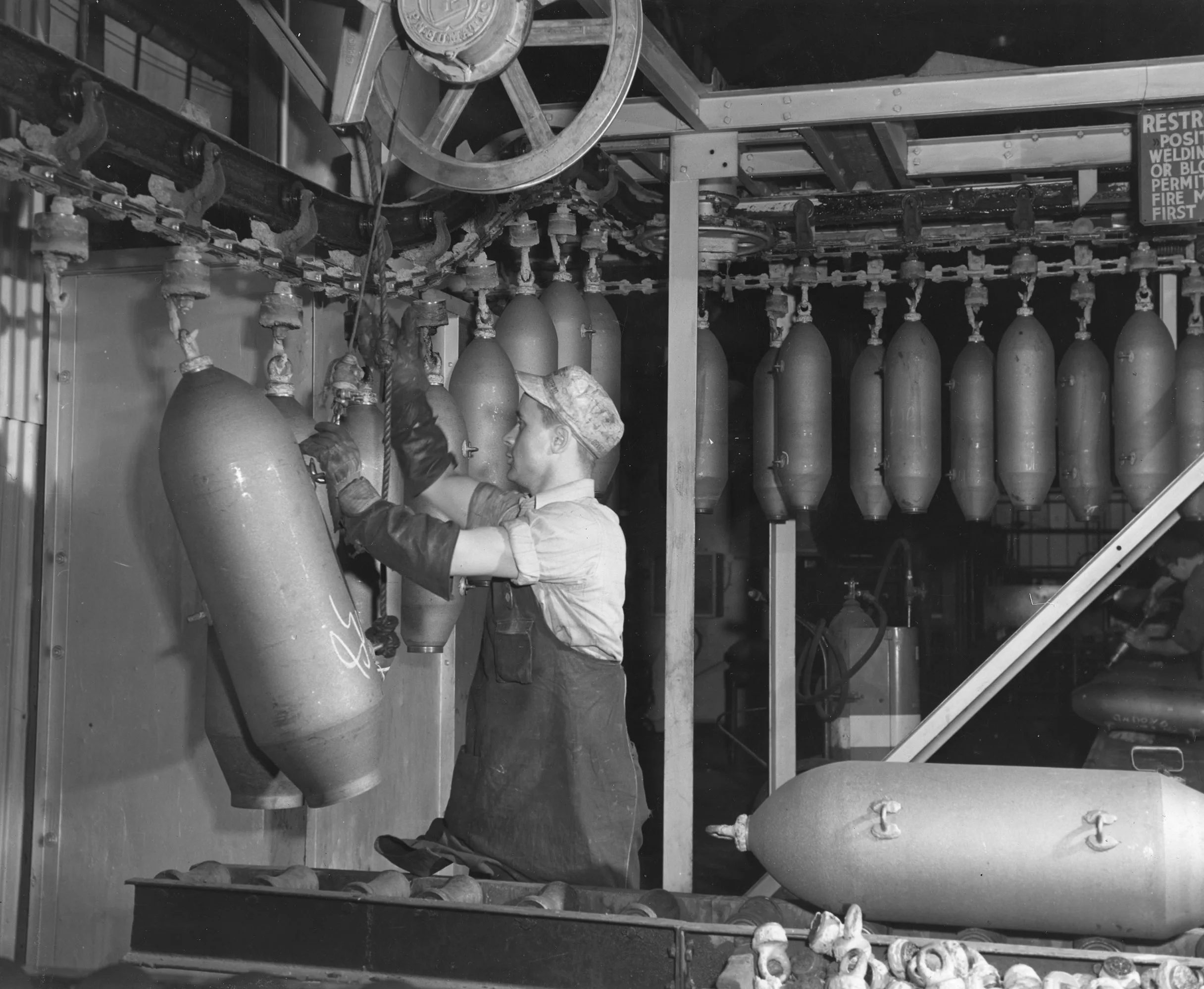 Factory worker in protective gear hangs large bomb casings onto an overhead conveyor system in a munitions plant.