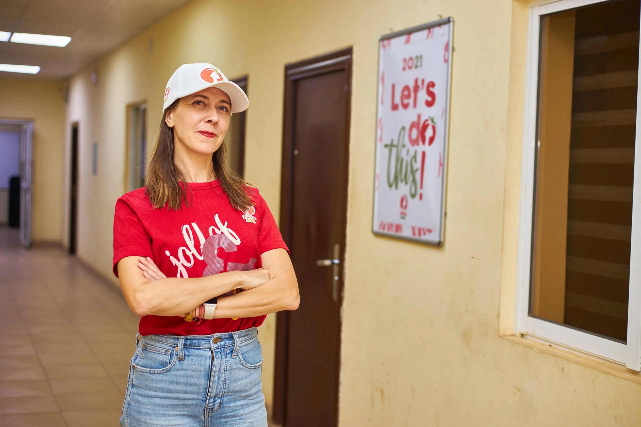 Person in a red "jollof" shirt and white cap stands with arms crossed beside a “Let’s do this!” poster in a hallway.