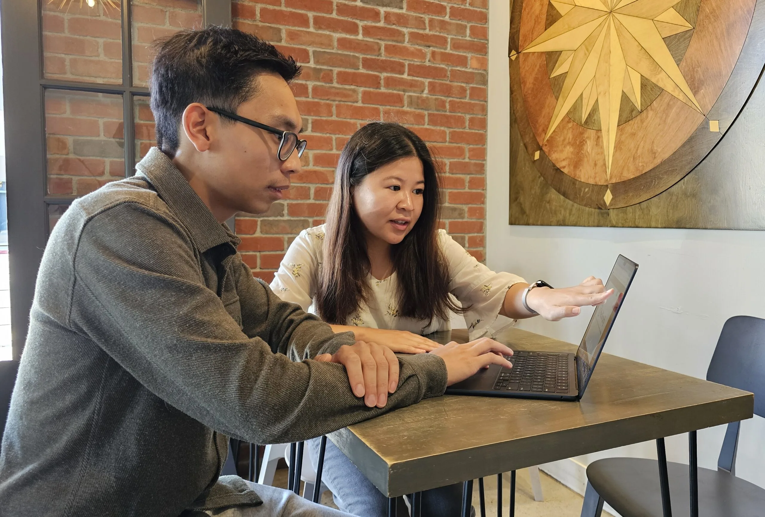 Two people sit at a table, with one pointing at a laptop screen while discussing something.