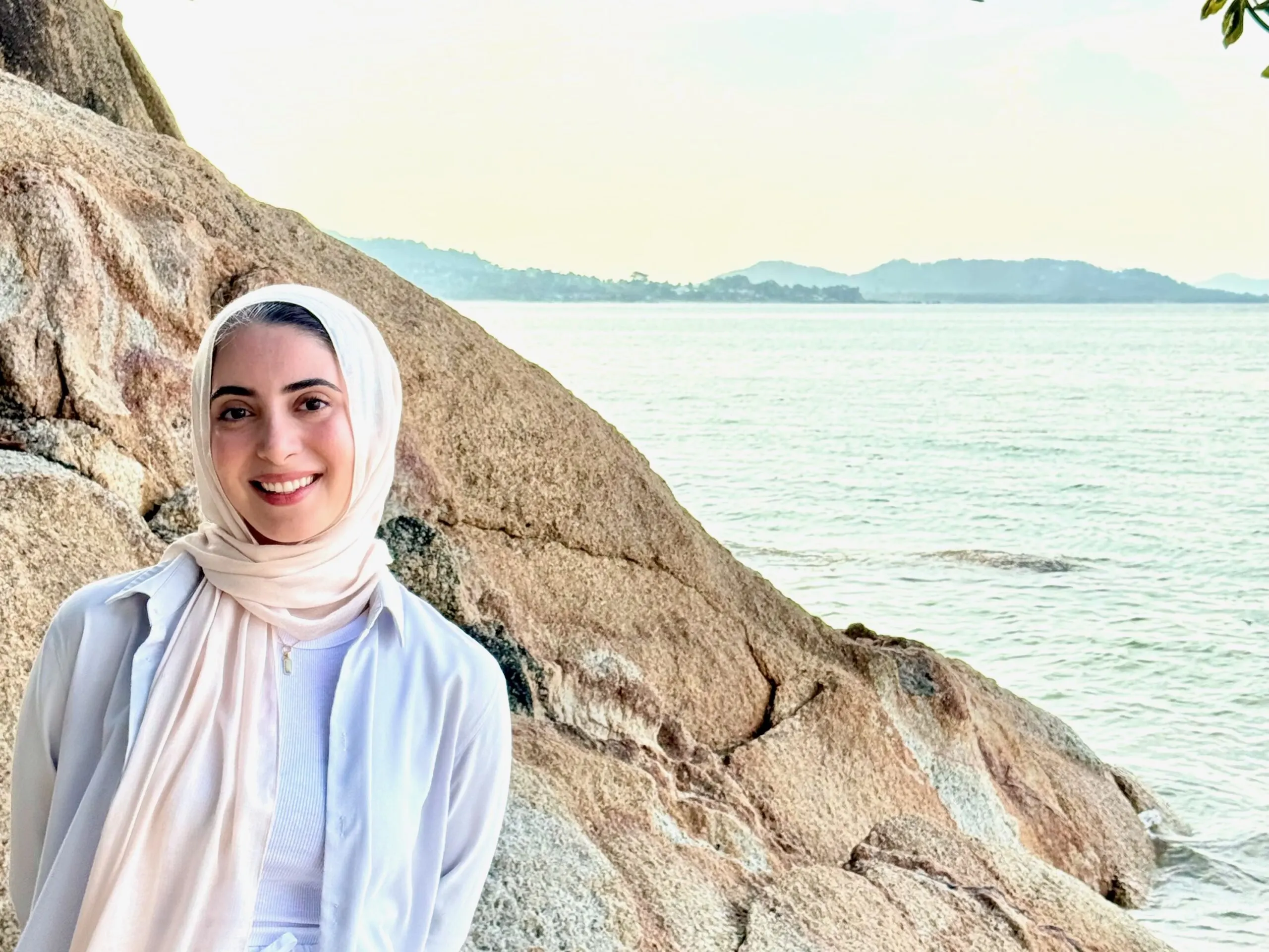 A young woman wearing a light pink hijab and white shirt smiles at the camera, standing by a large, textured rock formation on a beach. In the background, the ocean meets the sky with mountains or hills visible in the distance under hazy sunlight.