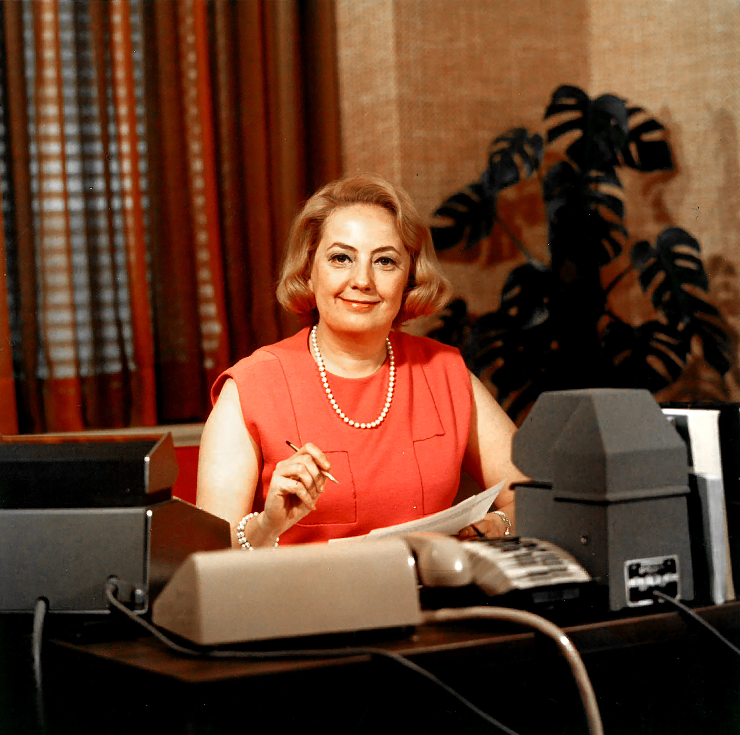 Muriel Siebert in sleeveless coral dress and pearls seated at a desk, holding papers, mid-20th-century office setting.