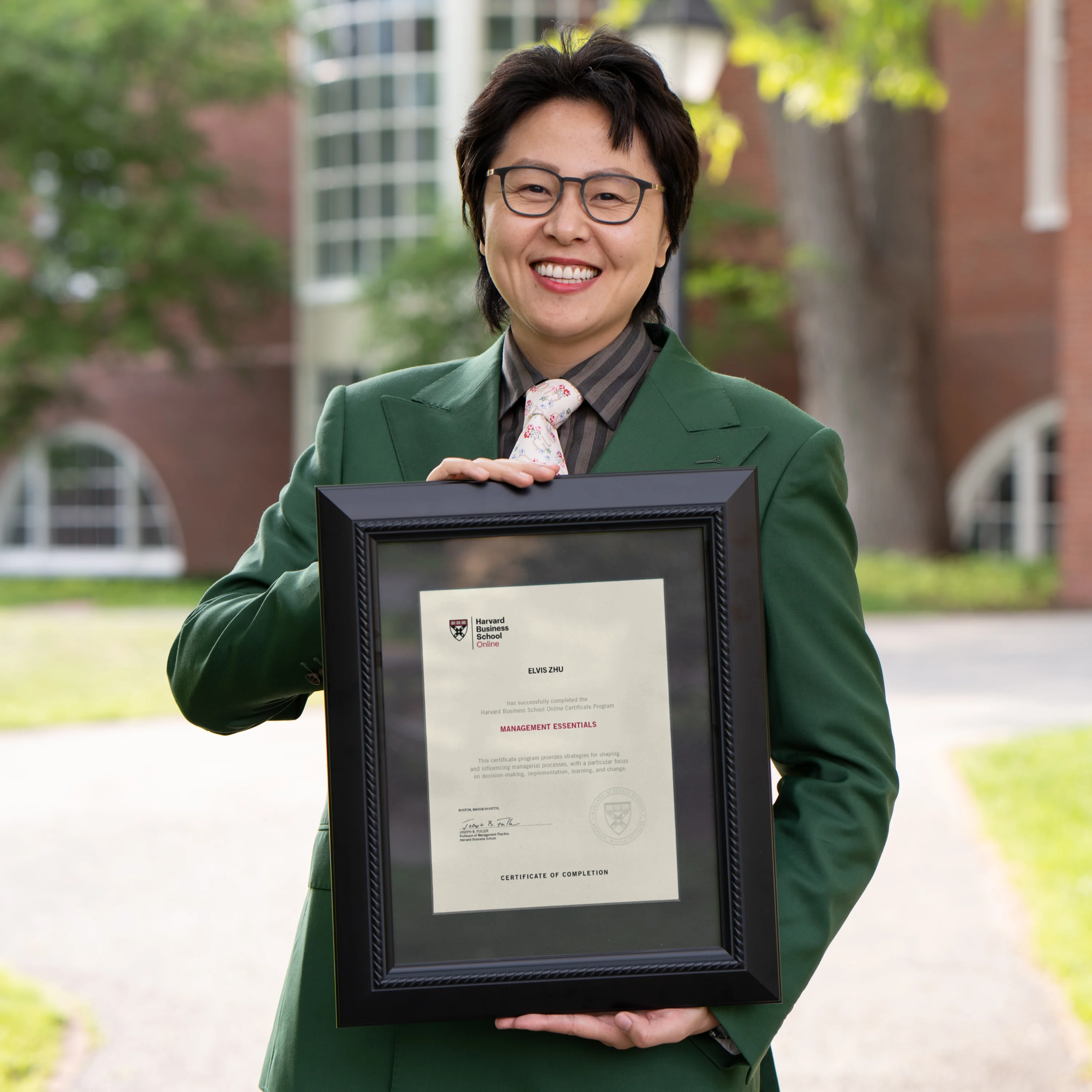 HBS Online community organizer in green suit smiling and holding a framed Harvard certificate outdoors.