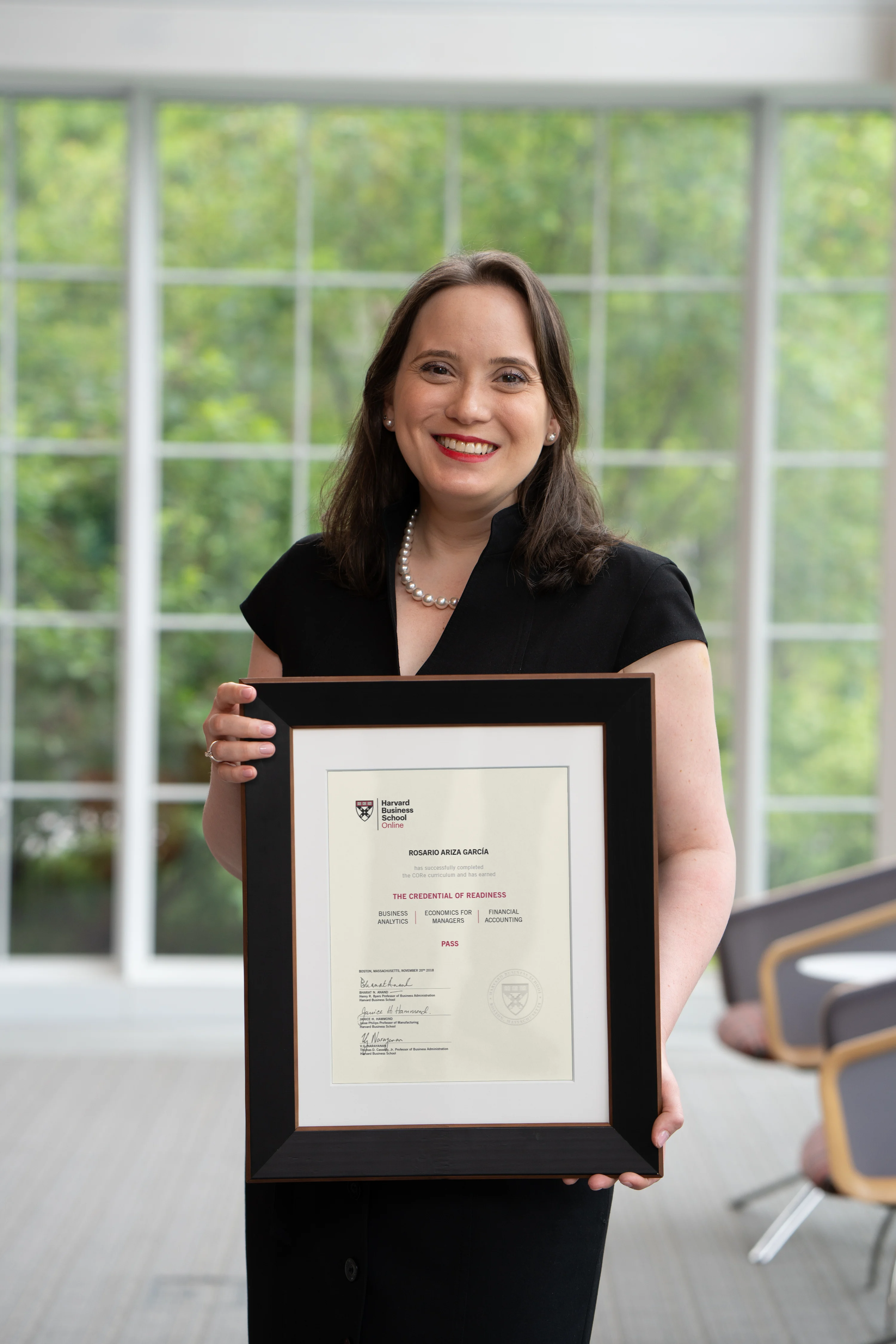 HBS Online community organizer in black dress smiling and holding holding a framed Harvard certificate in a bright room with large windows.