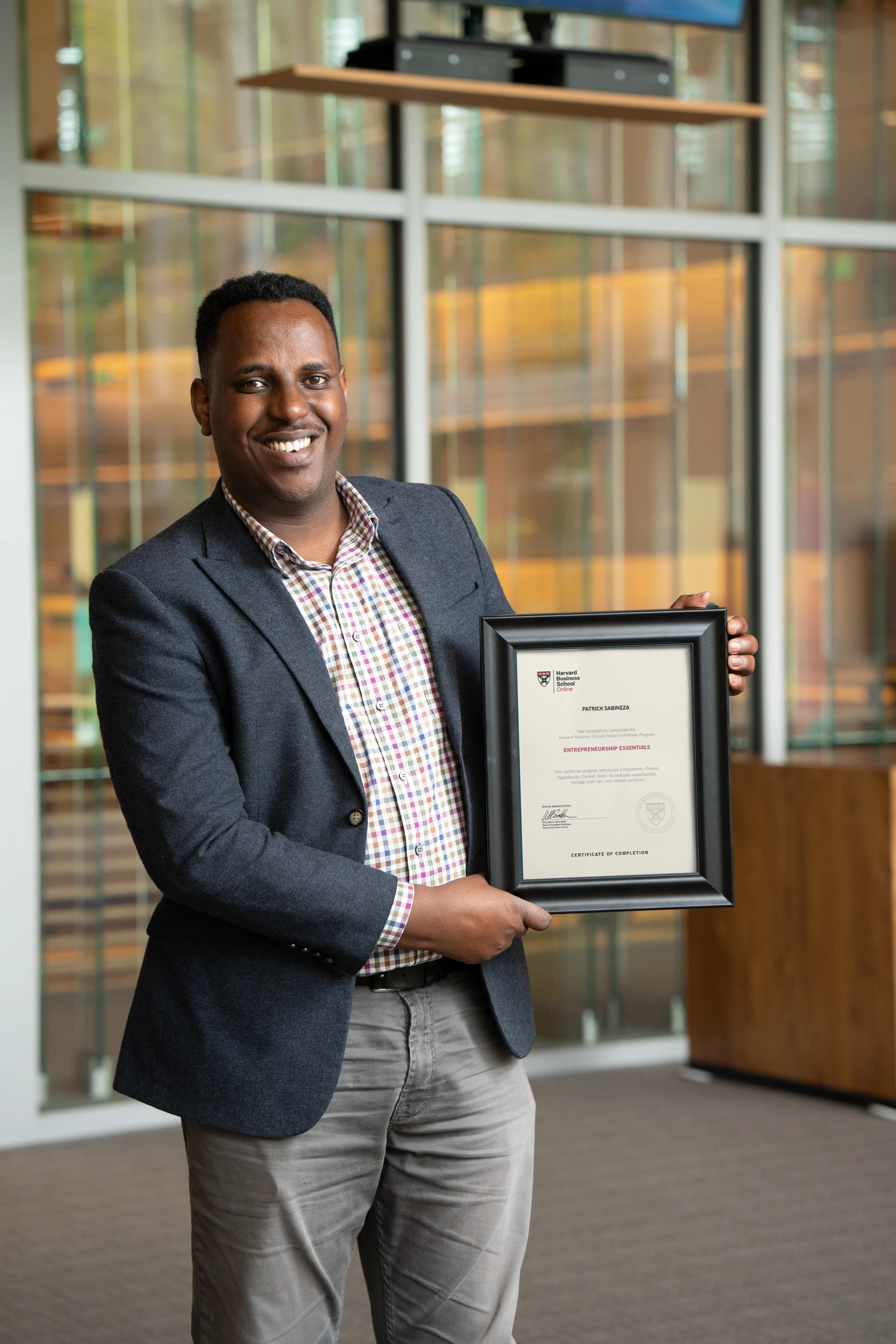 HBS Online community organizer in blazer smiling and holding a framed Harvard certificate indoors.