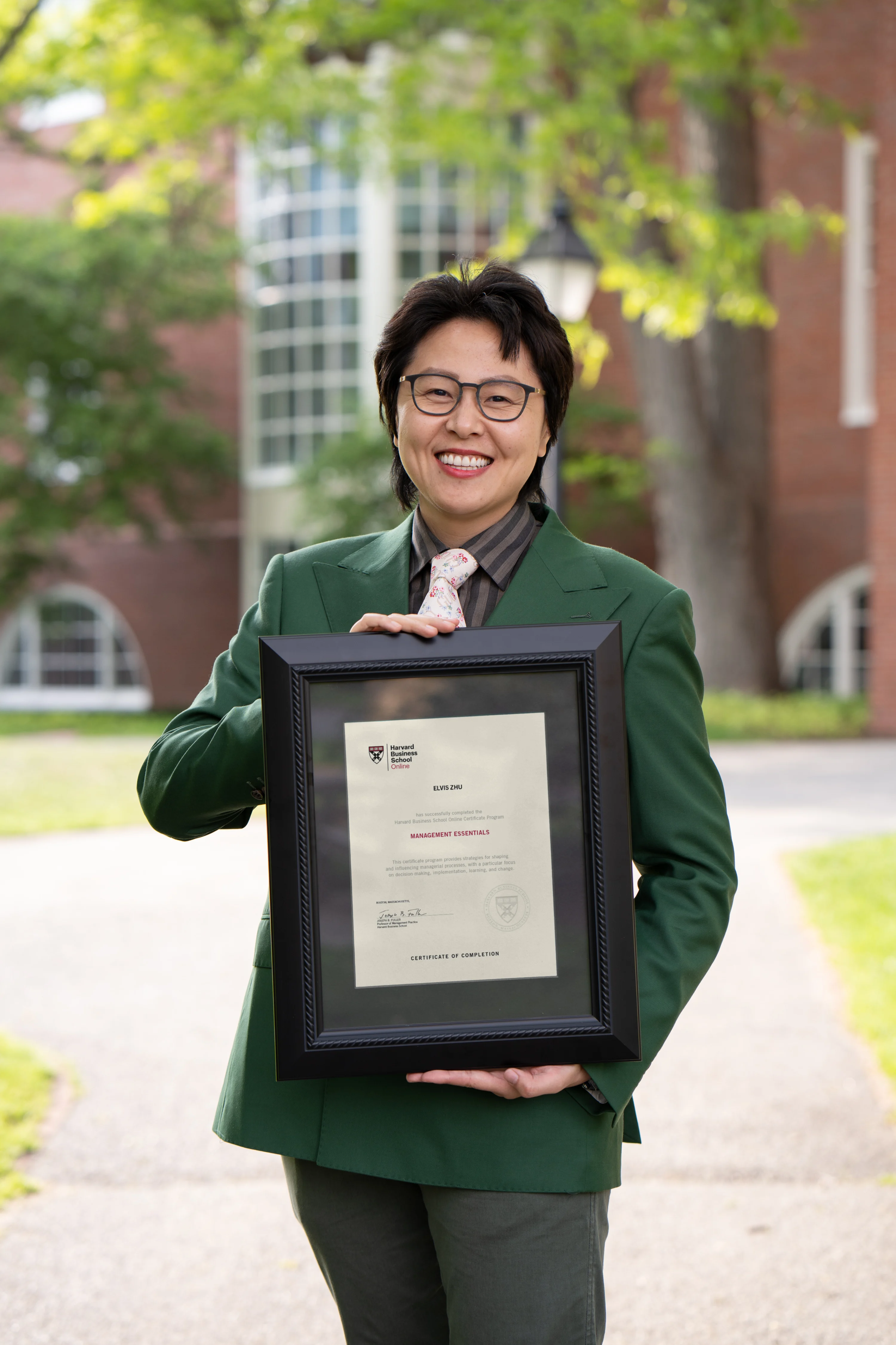 HBS Online community organizer in green suit smiling and holding a framed Harvard certificate outdoors.