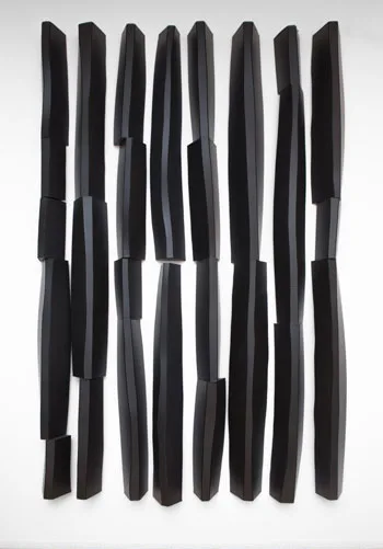 Wall-mounted sculpture of irregular vertical black forms made of corrugated plastic, arranged in twelve columns.