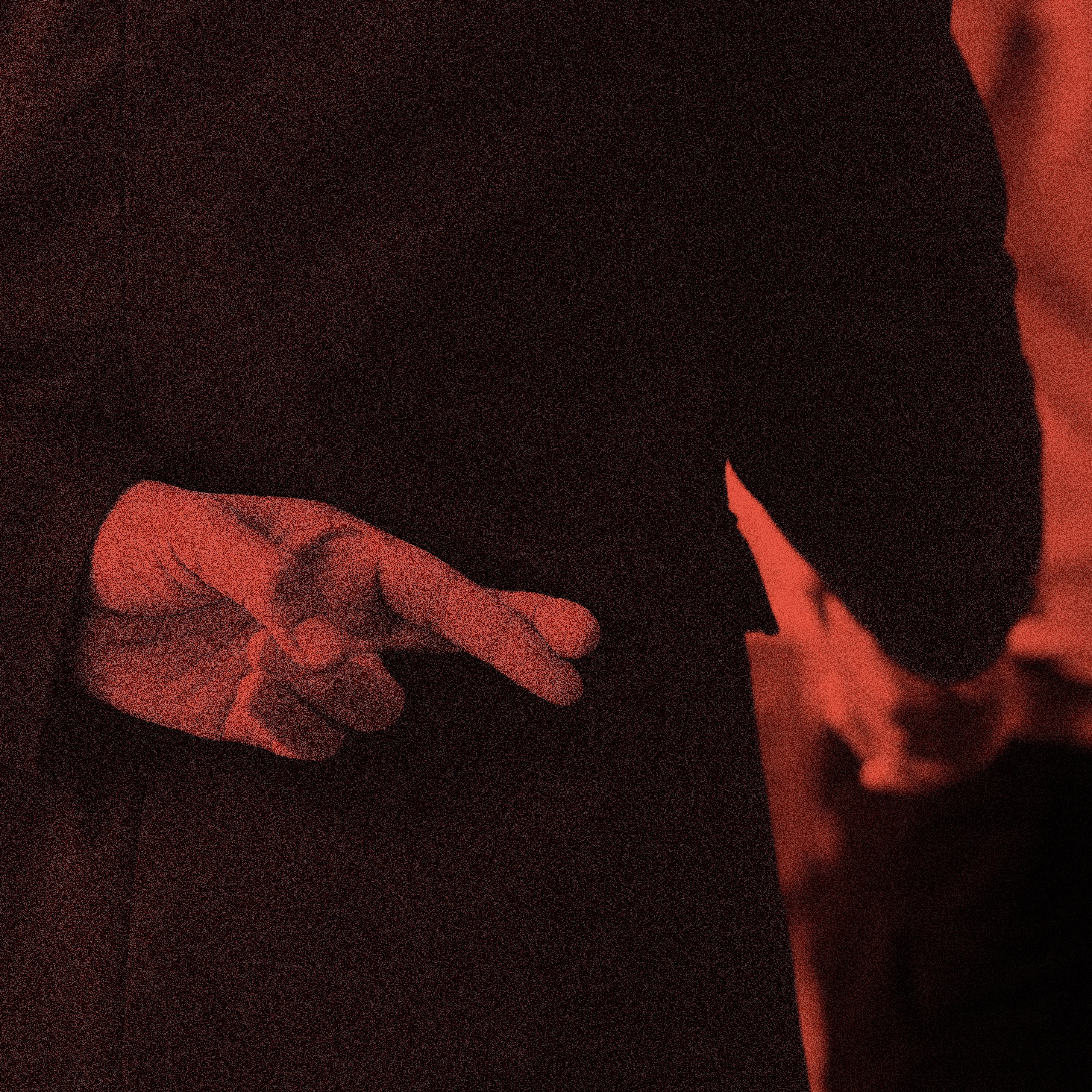 Close-up of a person’s hand behind their back with two fingers crossed, while the other arm extends forward out of frame, suggesting a handshake. The image is tinted red.