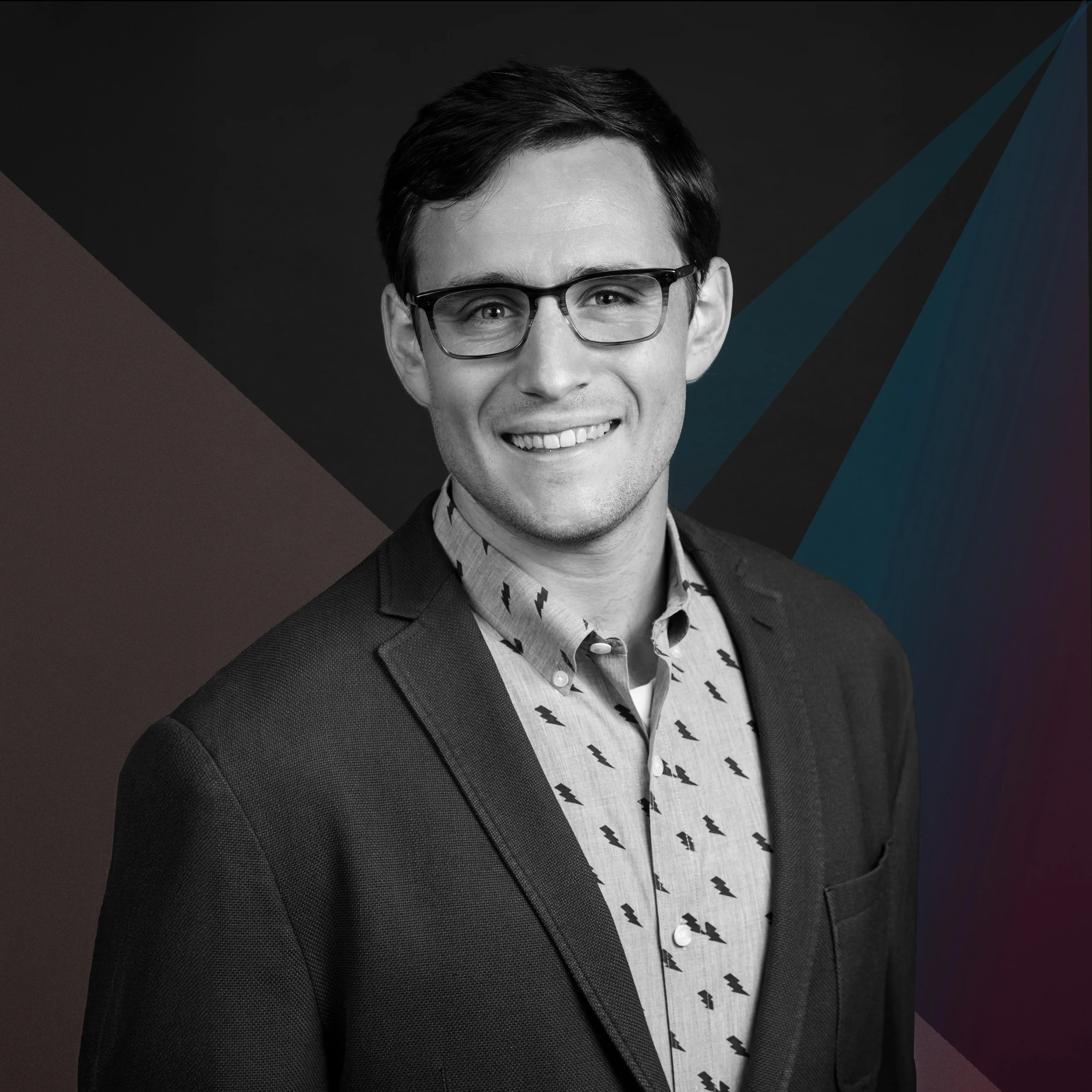 A smiling man wearing glasses, a patterned shirt, and a blazer. The image is in black and white with subtle colored accents behind him.