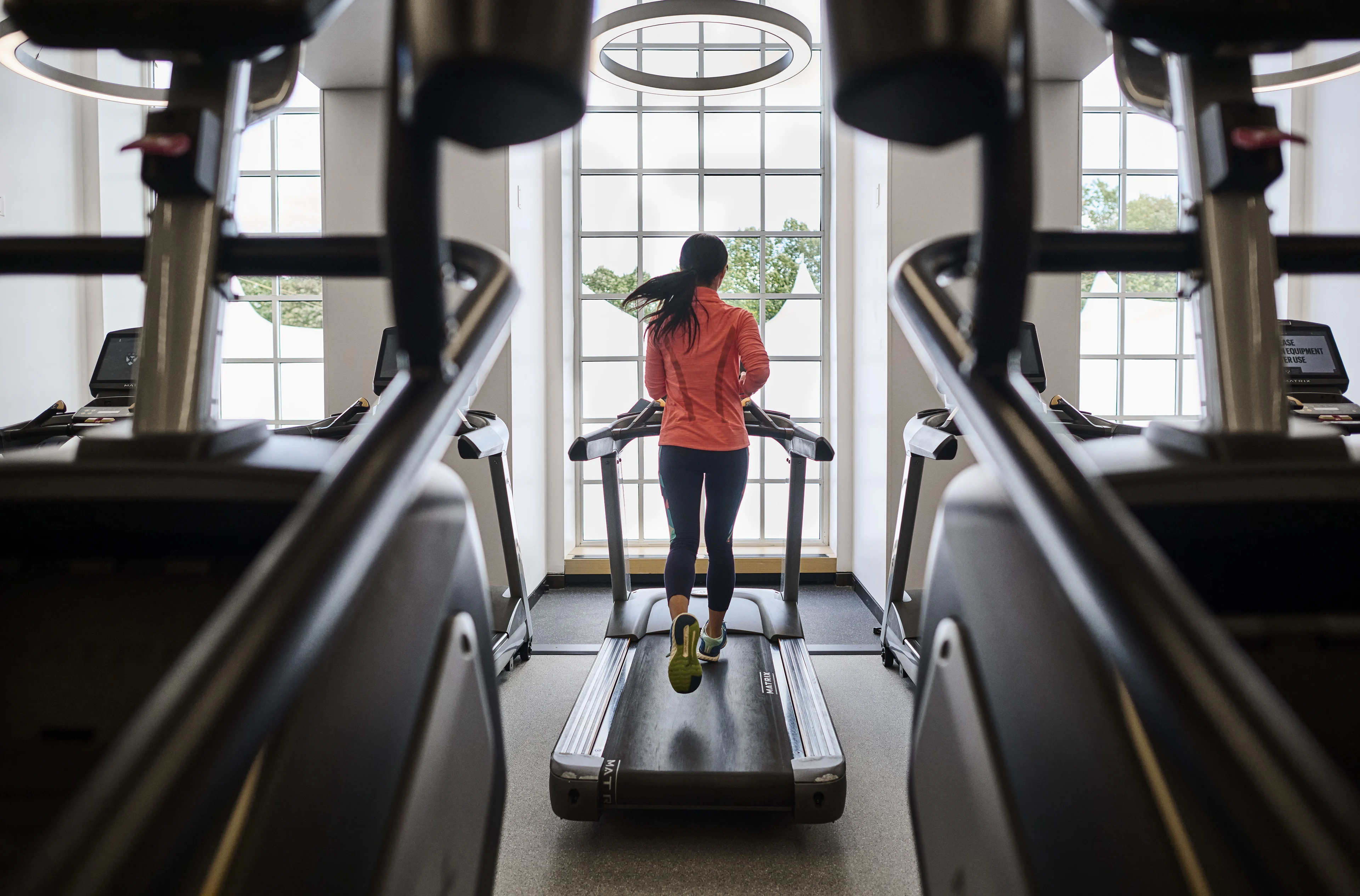 A woman runs on a treadmill in front of a large window wearing a peach colored top and black leggings.