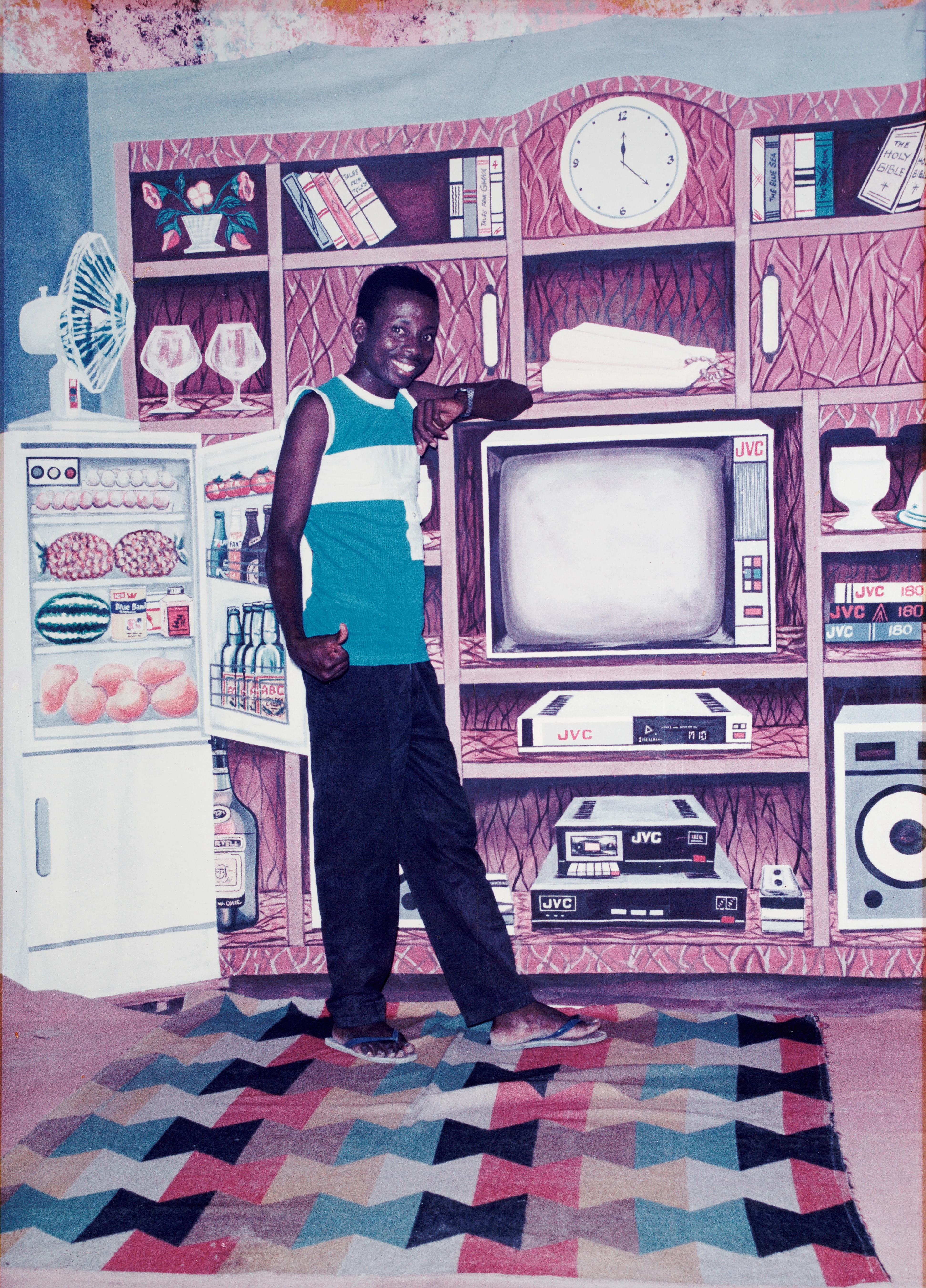 A smiling boy poses before a painted backdrop of a stocked fridge, electronics, and luxury furnishings.