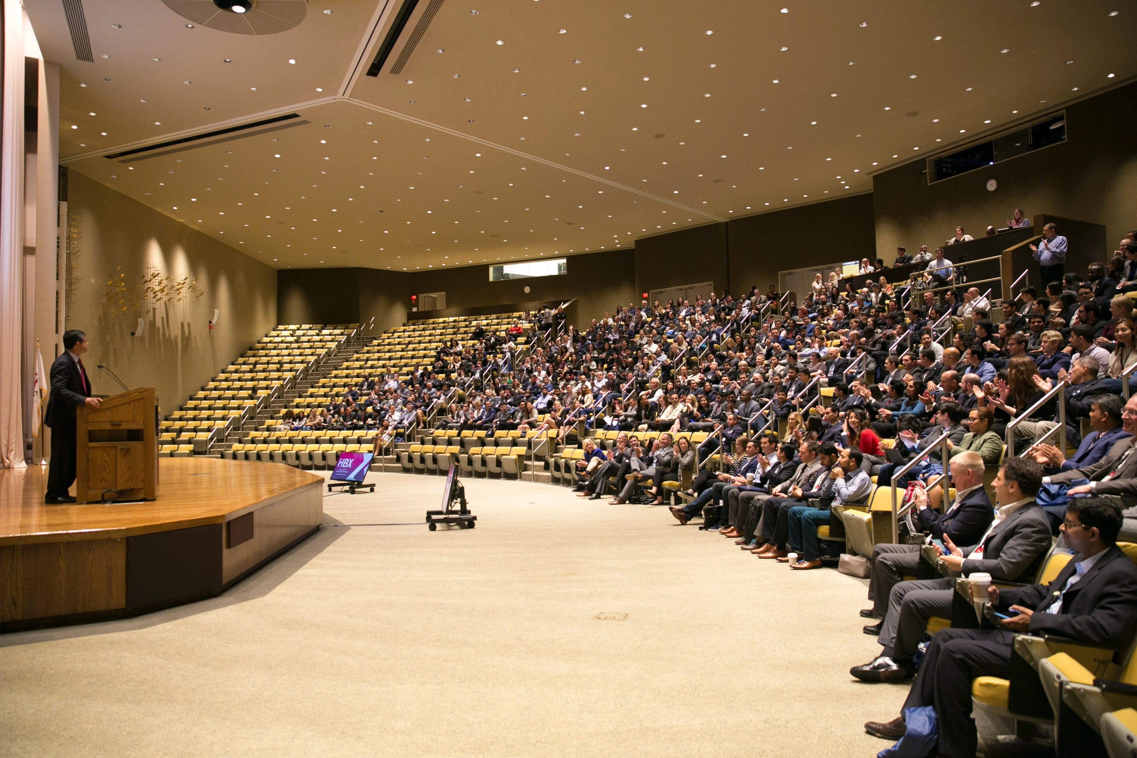 Professor Bharat Anand stands at the podium on stage and welcomes an auditorium full of students to HBX ConneXt