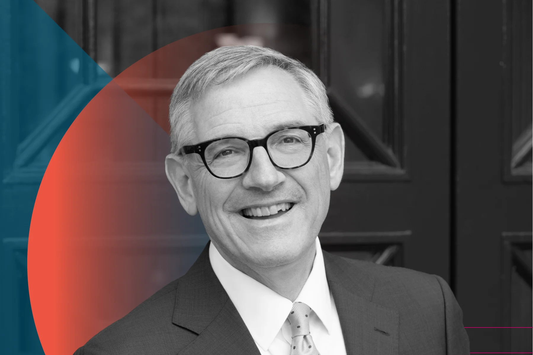 A black and white portrait of a smiling person with short hair, wearing glasses, a dark suit, and a light polka-dot tie, standing in front of dark double doors with a blue and red geometric overlay.