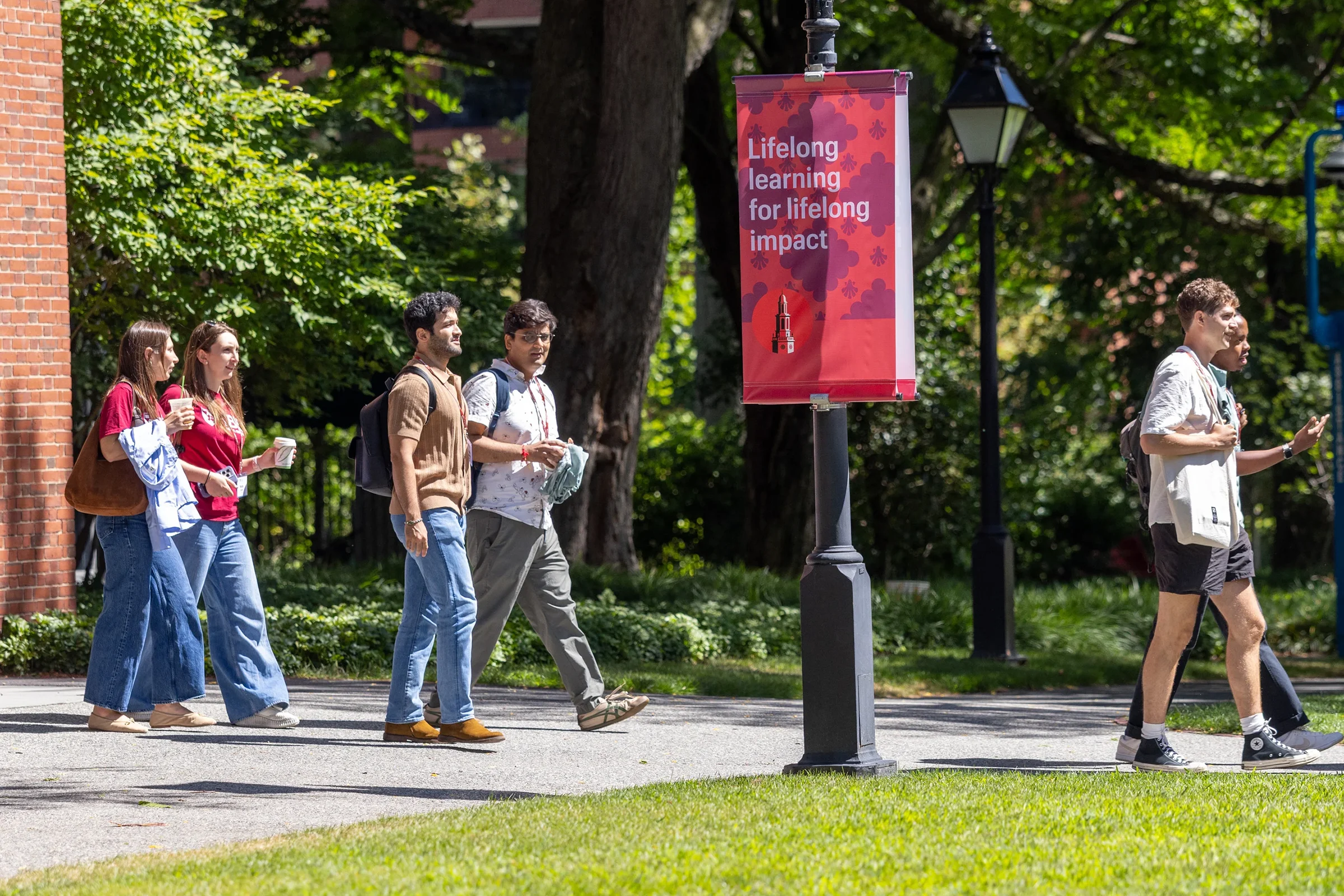 Students walking on the Harvard campus near a banner reading “Lifelong learning for lifelong impact.”