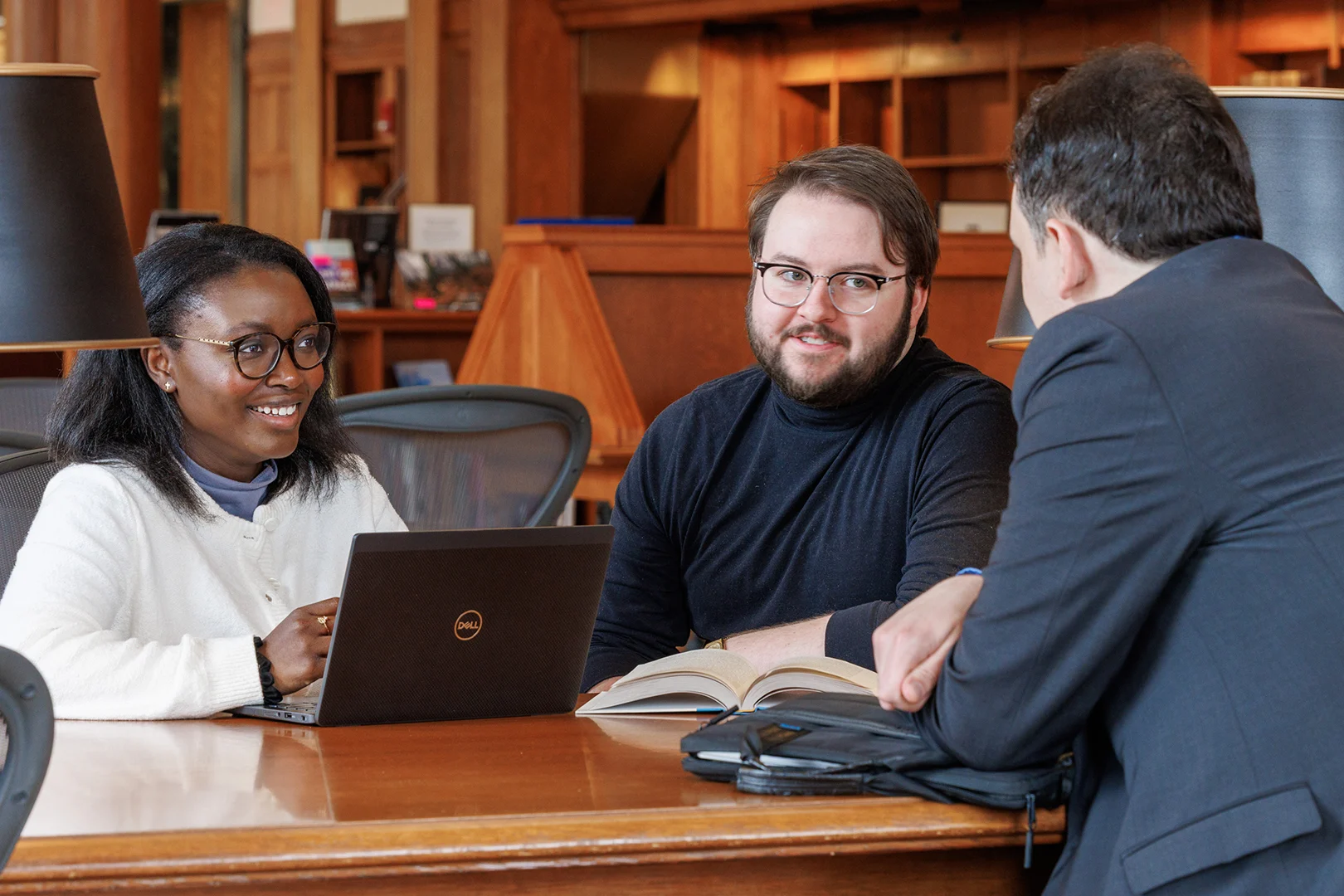 Three people having a discussion at a wooden table with a laptop and open book in Baker Library.