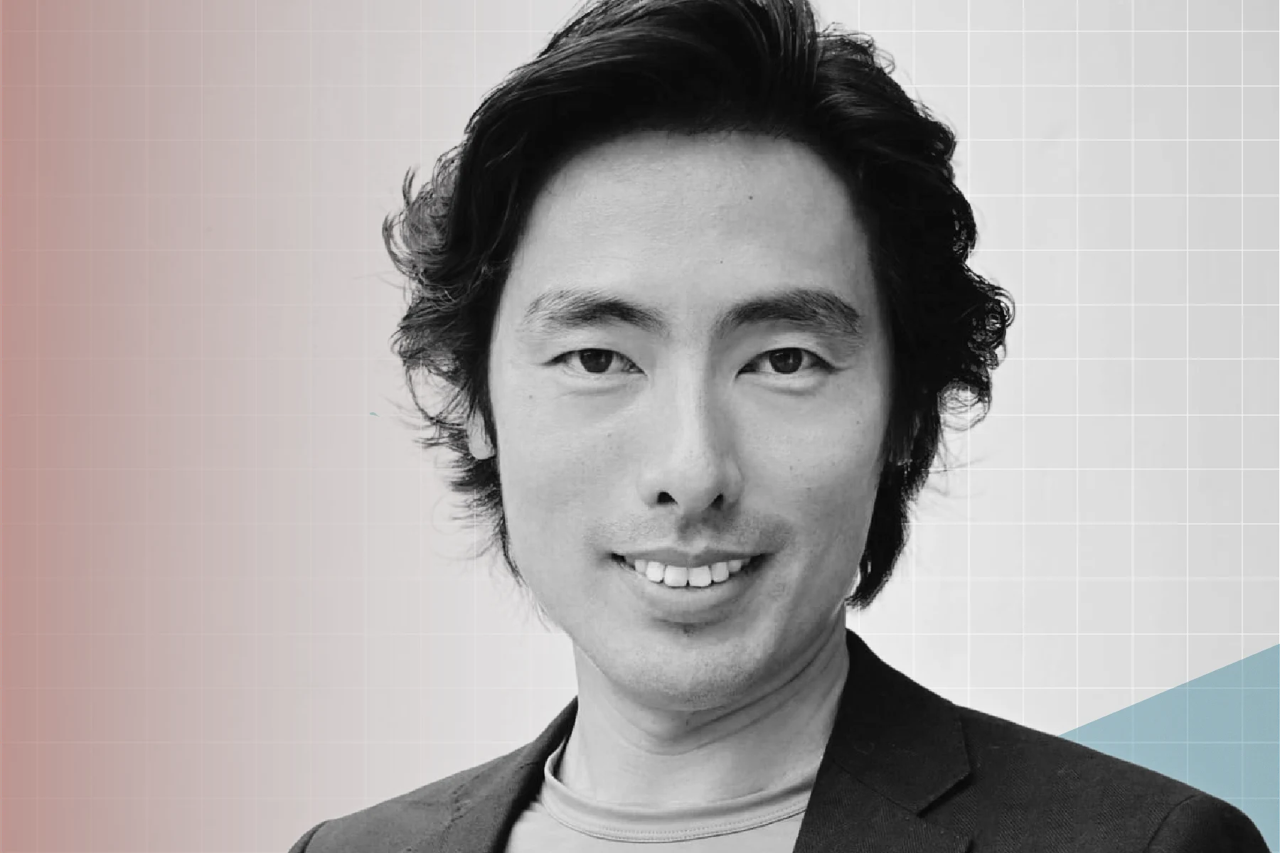 Person with medium-length, slightly wavy dark hair and they are smiling and looking directly at the viewer, wearing a dark blazer over a light-colored crew neck shirt, with a pale, geometric pattern in the background.