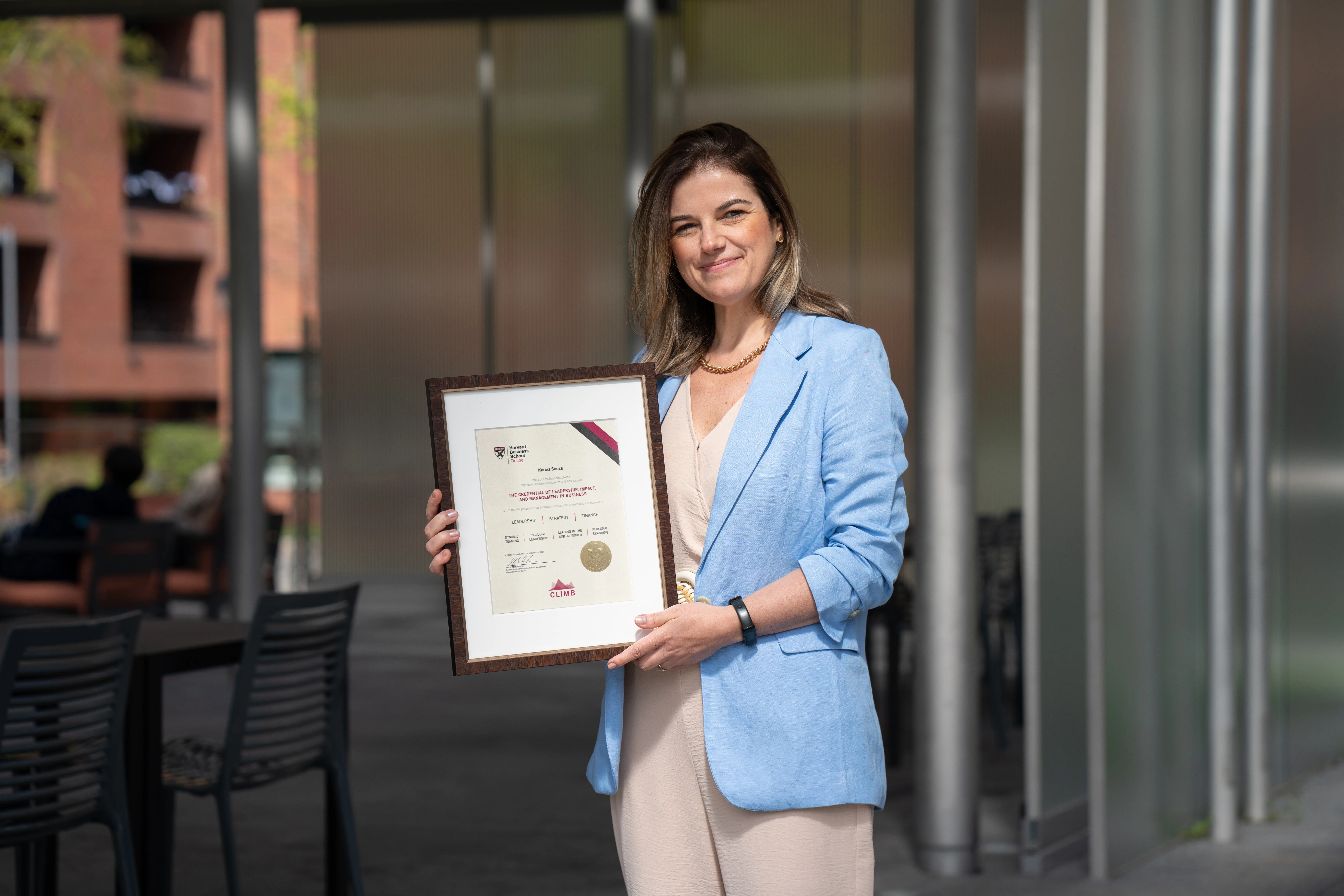 Individual holding a Harvard certificate of achievement, smiling outside a modern campus building.