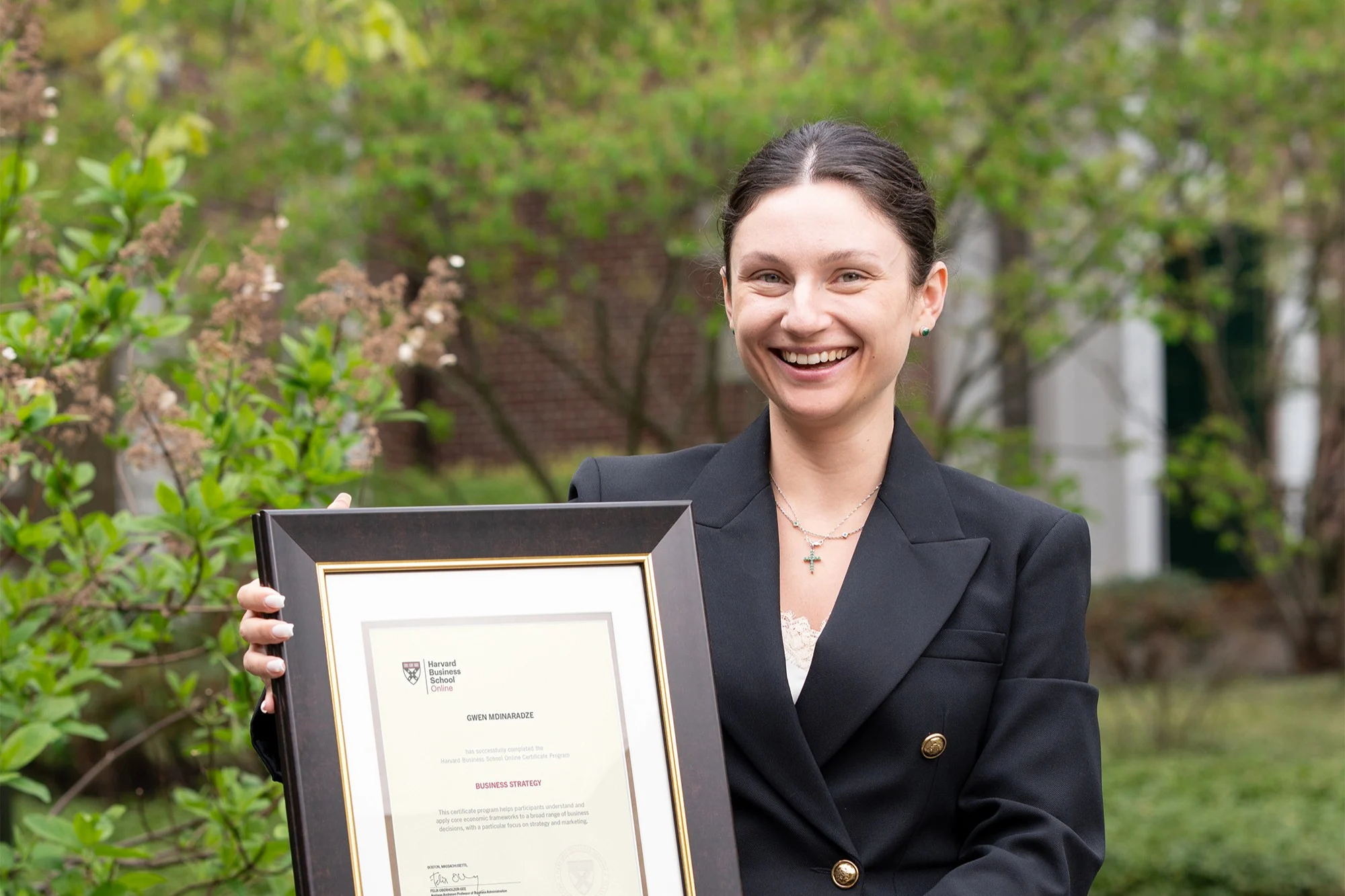 HBS Online community organizer in black suit smiling and holding a framed Harvard certificate outdoors.