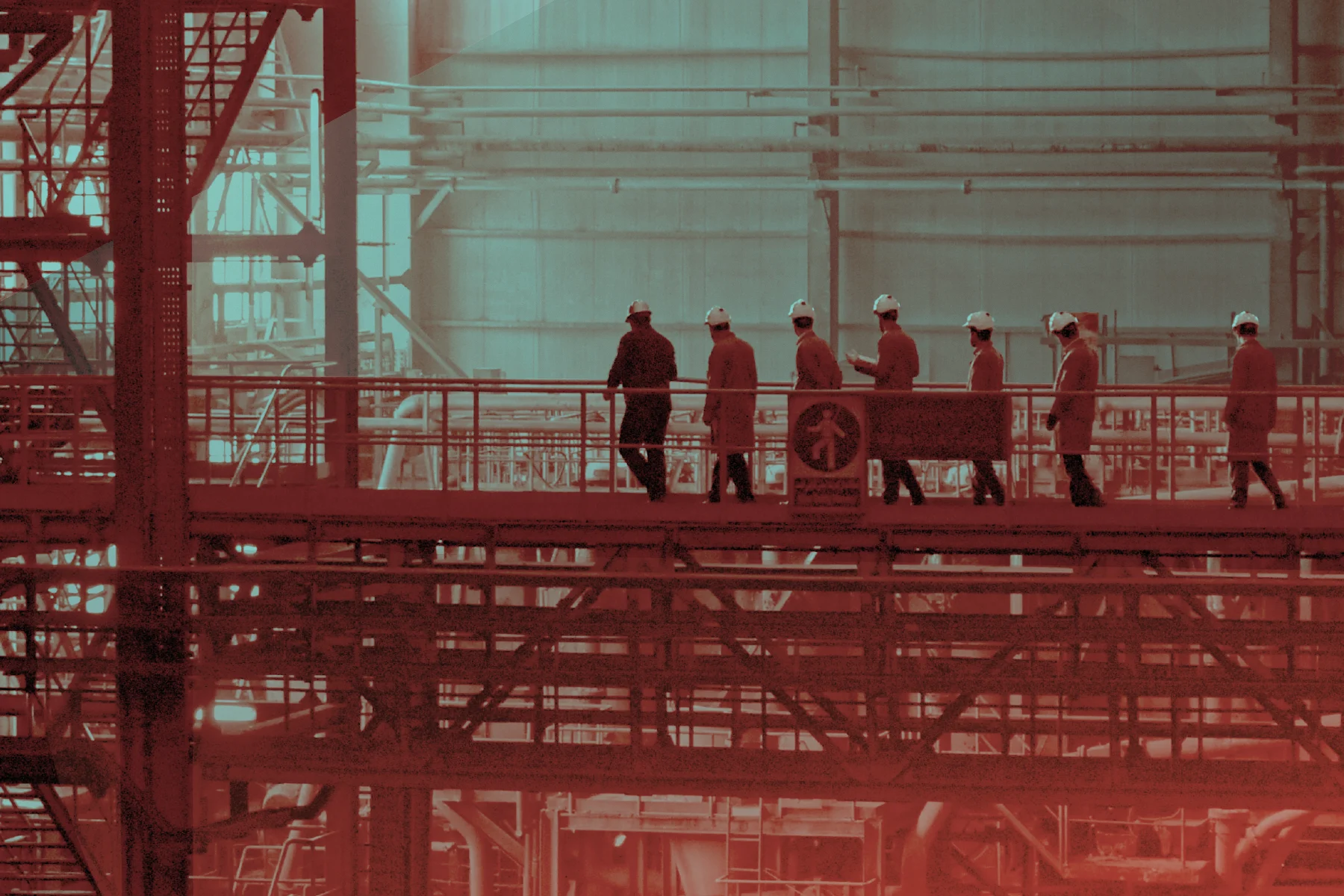 Workers in hard hats walk across an elevated platform inside a large industrial facility with metal beams, pipes, and tall windows, tinted in red and teal.