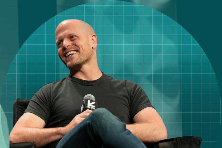 Tim Ferriss sitting on a dark armchair, holding a microphone. He wears a fitted dark T-shirt, blue jeans, and light-colored lace-up shoes. His legs are crossed, and he's turned slightly. Behind him is a teal circular backdrop with a grid pattern.