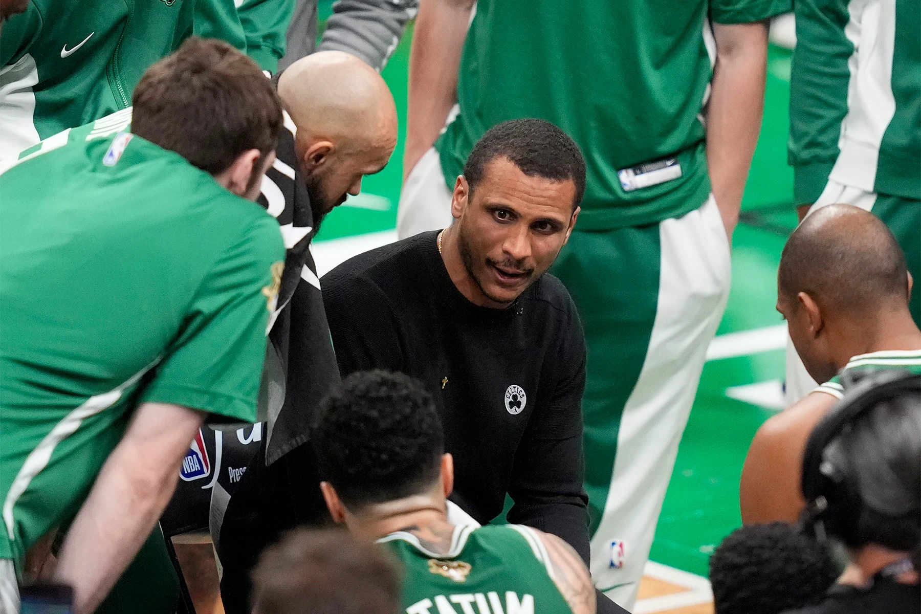 Joe Mazzulla, in a black shirt is seated, surrounded by men in green and white basketball uniforms.