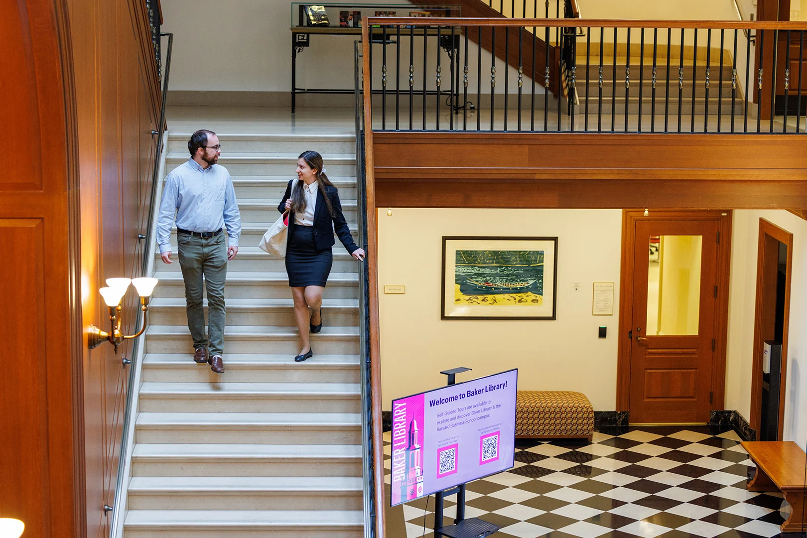 Two people walk down a staircase inside Baker Library, passing a digital welcome sign in a well-lit, wood-accented interior.