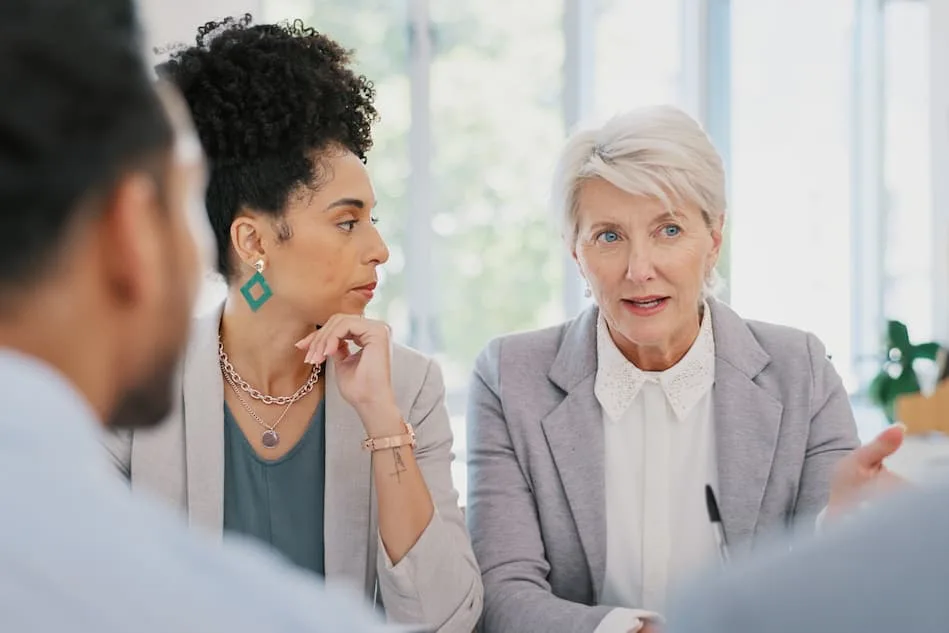 Two colleagues listen during a professional meeting, with one person speaking beside another with two attendees in the foreground.