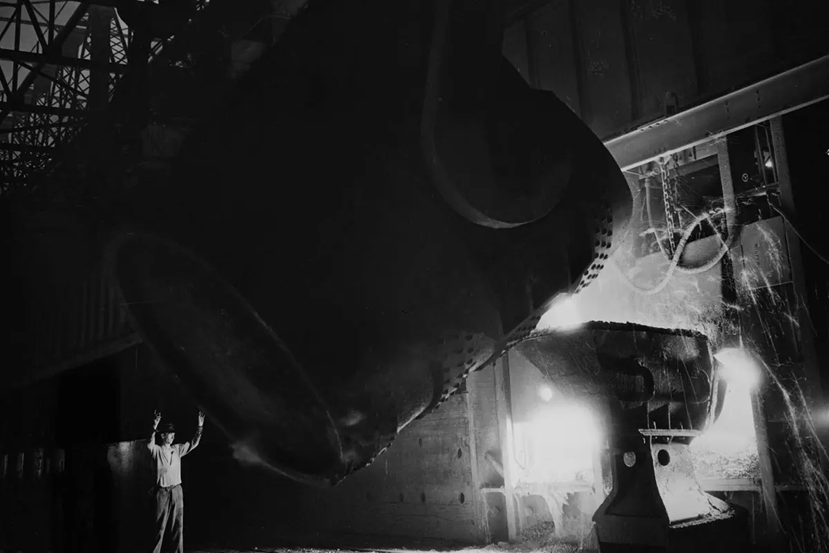 Worker guides massive ladle pouring molten metal into a mold at a dimly lit steel mill.