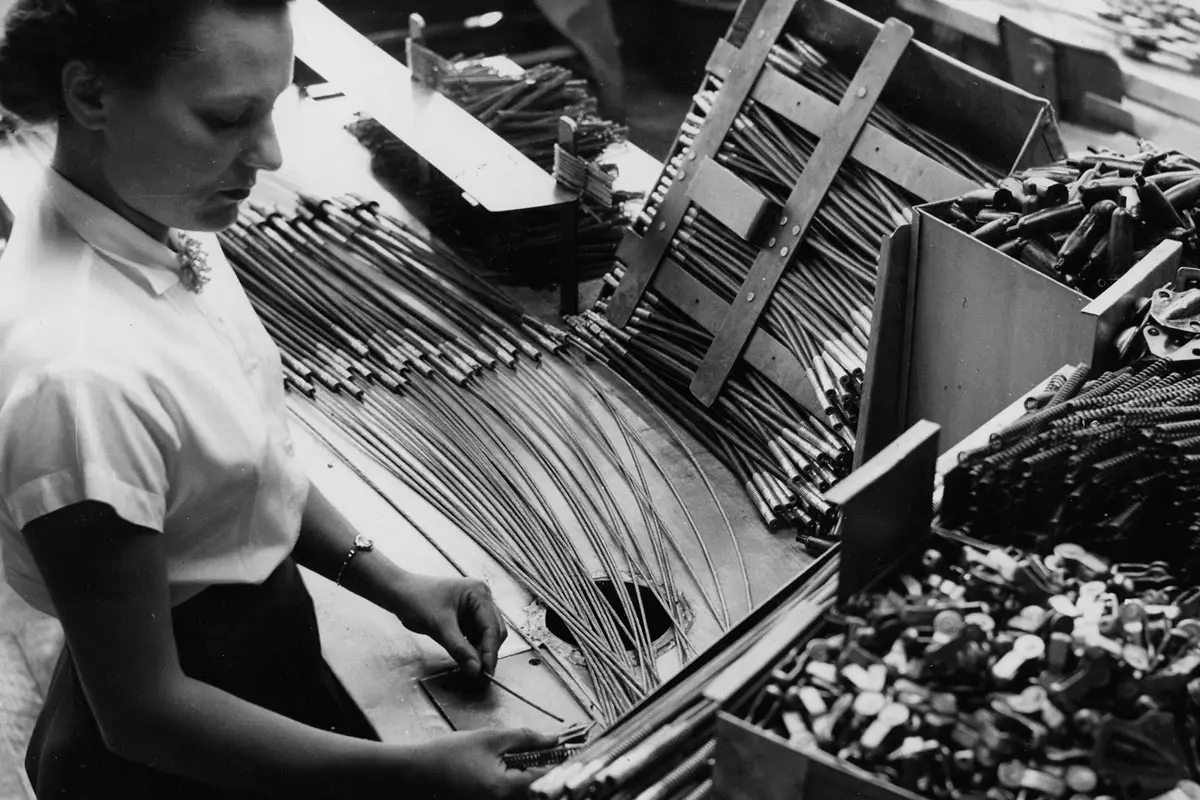 Factory worker organizes metal rods and components on an assembly table surrounded by bins.