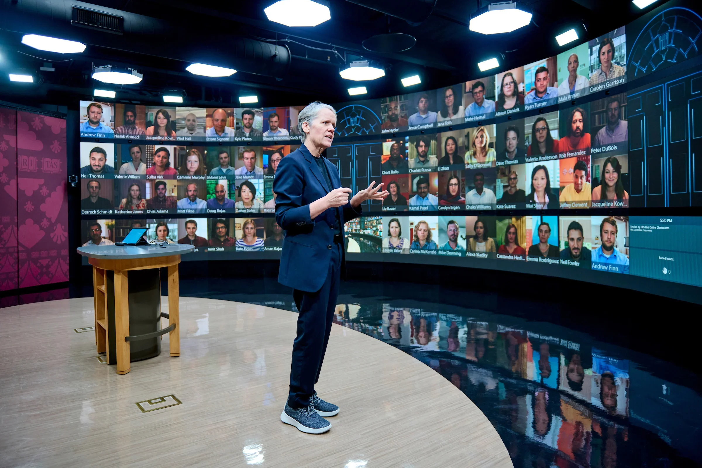 Instructor gestures in a virtual classroom with a large video wall of participants and a digital chalkboard