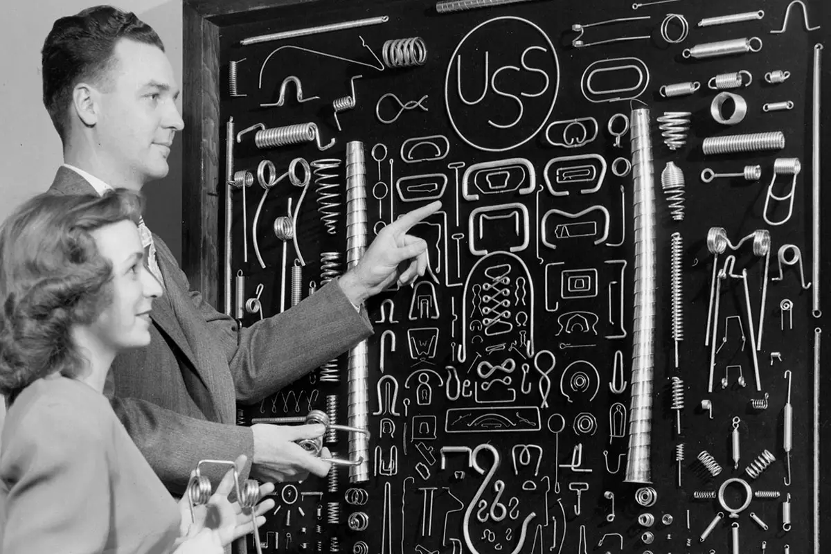 Two individuals examine a display board of various metal springs and wire forms labeled with the USS logo.